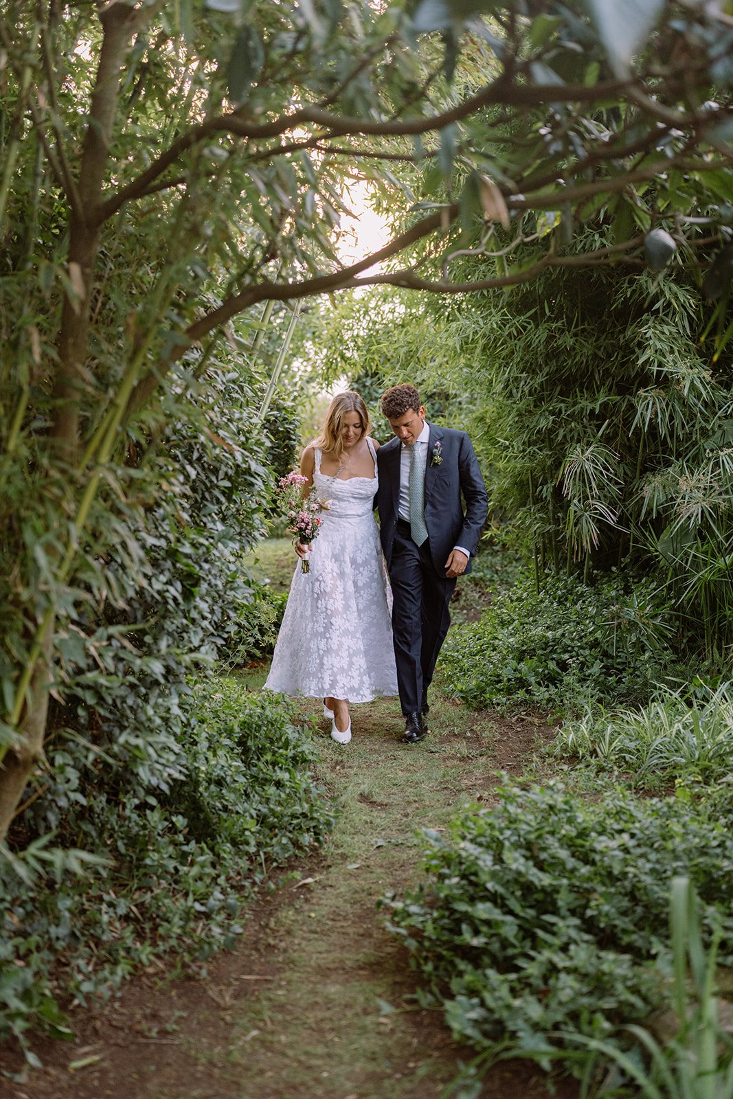 A bride and groom walk together on a narrow garden path surrounded by lush green plants and trees.