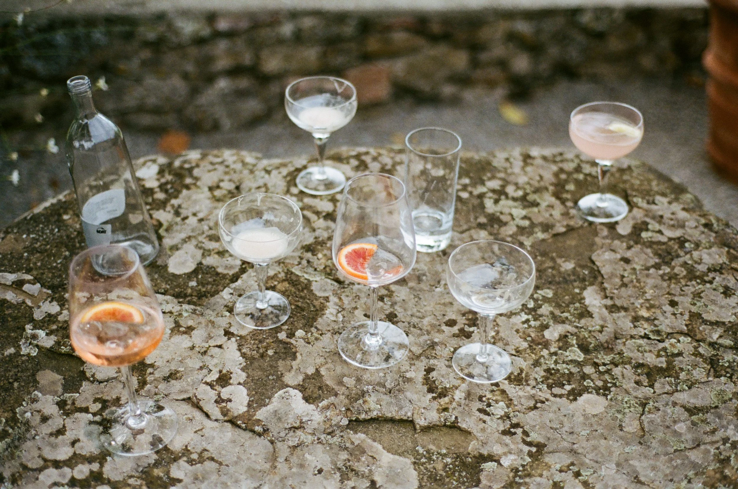 Assorted cocktail glasses on a weathered stone table outdoors, some with slices of citrus fruit, and one with a slice of grapefruit.