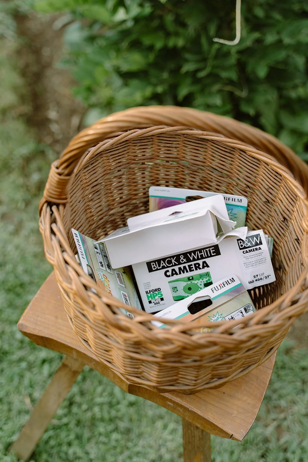 A woven wicker basket filled with camera film packages sits on a small wooden stool outdoors, with greenery in the background.