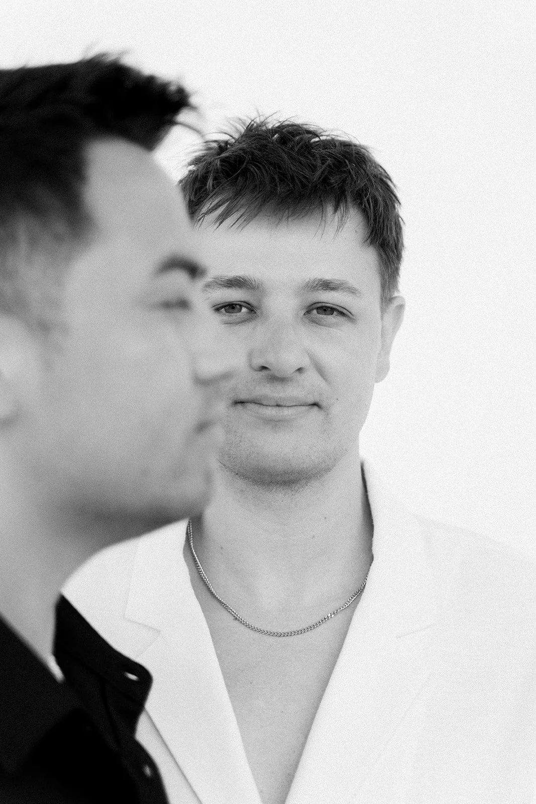 Black and white photograph of two young men, one in focus with short wavy hair, smiling subtly, wearing a white coat and a chain necklace, the other out of focus with short dark hair, wearing a dark shirt.
