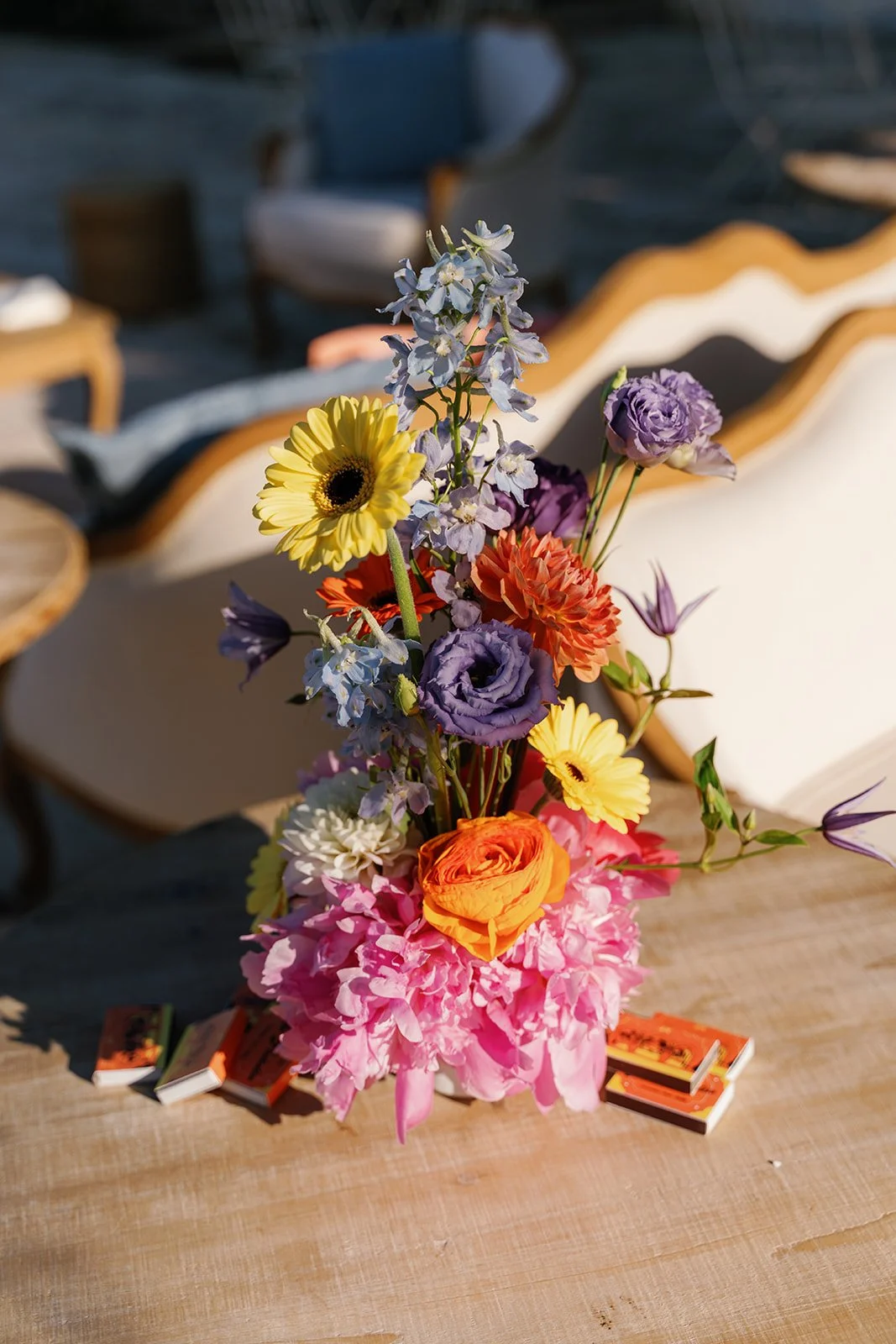 A colorful flower bouquet with yellow, purple, orange, pink, and white flowers on a wooden table, with a blurred outdoor patio background.