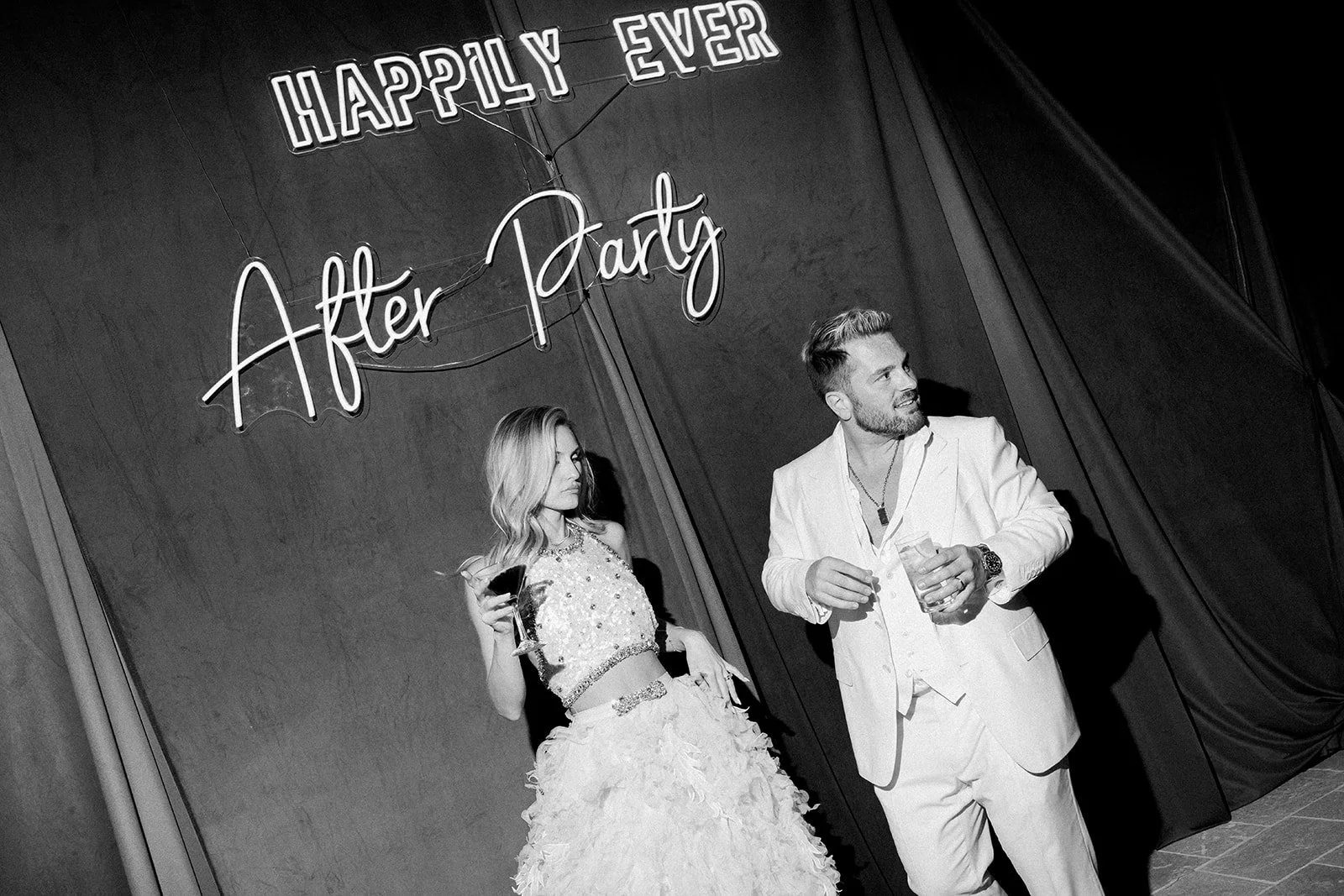 A black and white photo of a man and woman at a party with a sign that says 'Happily Ever After Party' in the background. The woman is wearing a fancy dress and holding a drink, while the man is dressed in a white suit jacket and also holding a drink