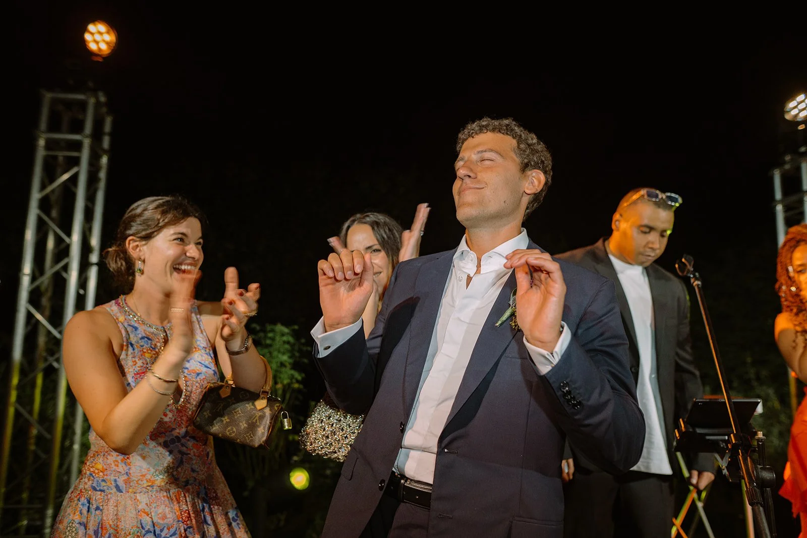 A man in a dark suit and white shirt dancing with a smile at an outdoor night event, surrounded by smiling women and a man in the background, under stage lights.