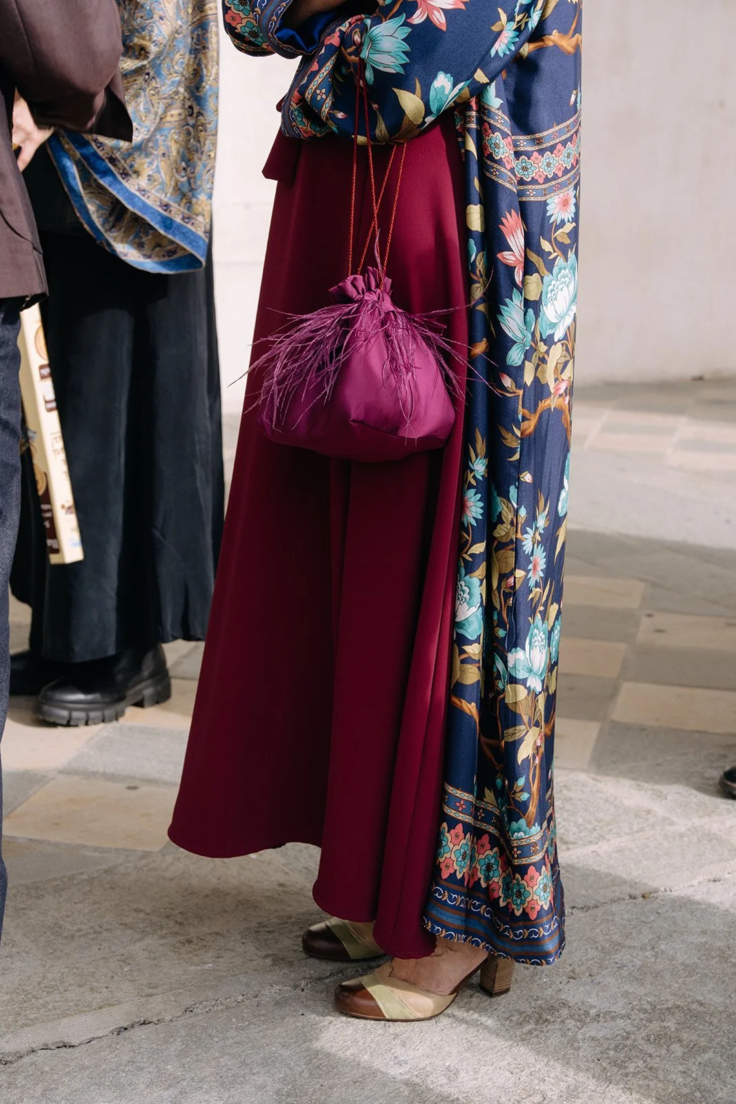 Close-up of a person wearing colorful patterned pants and a floral blouse, holding a small purple bag with feather details, standing on a tiled floor.