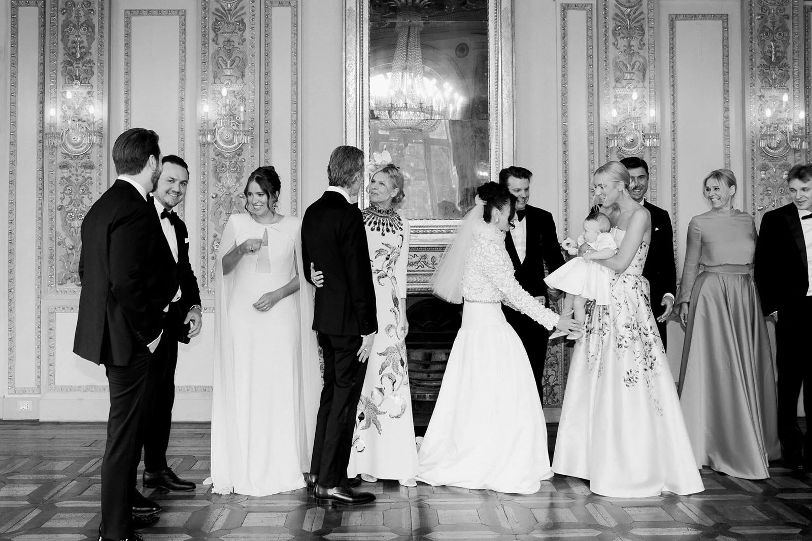 A black and white photo of a wedding ceremony with people dressed in formal attire, gathered in an ornate room with decorative walls and a chandelier reflected in a mirror.