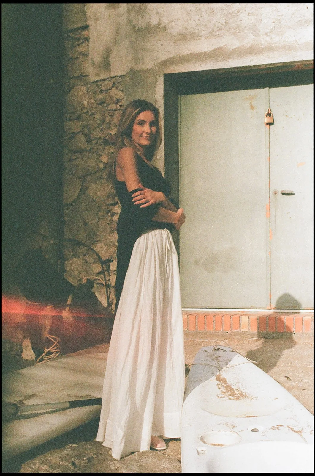 A woman in a black top and white wide-legged pants stands outdoors against a stone wall and door, with construction or renovation materials around her.