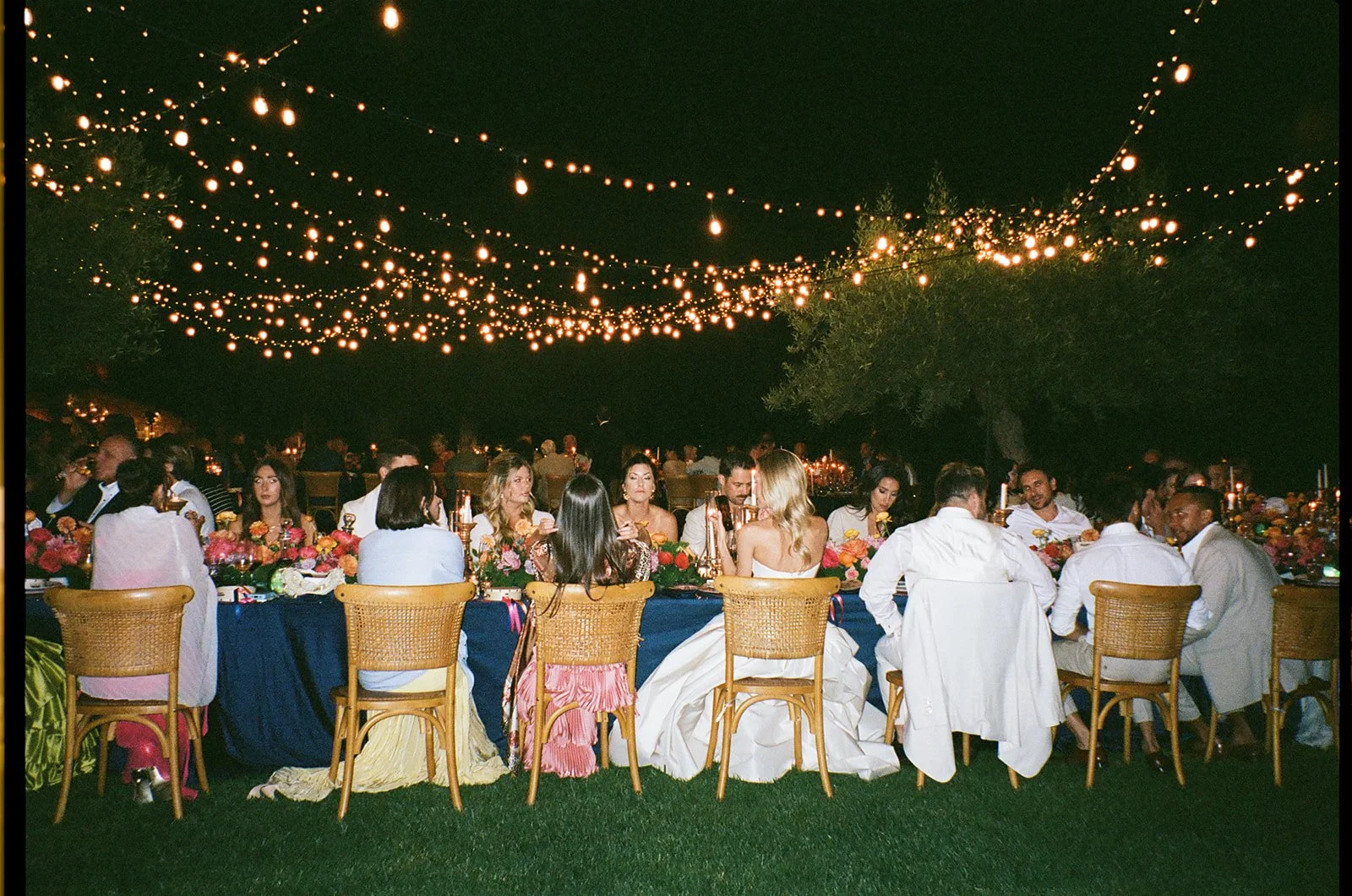 An outdoor evening dinner party with people seated at a long table under string lights, with floral centerpieces and greenery.