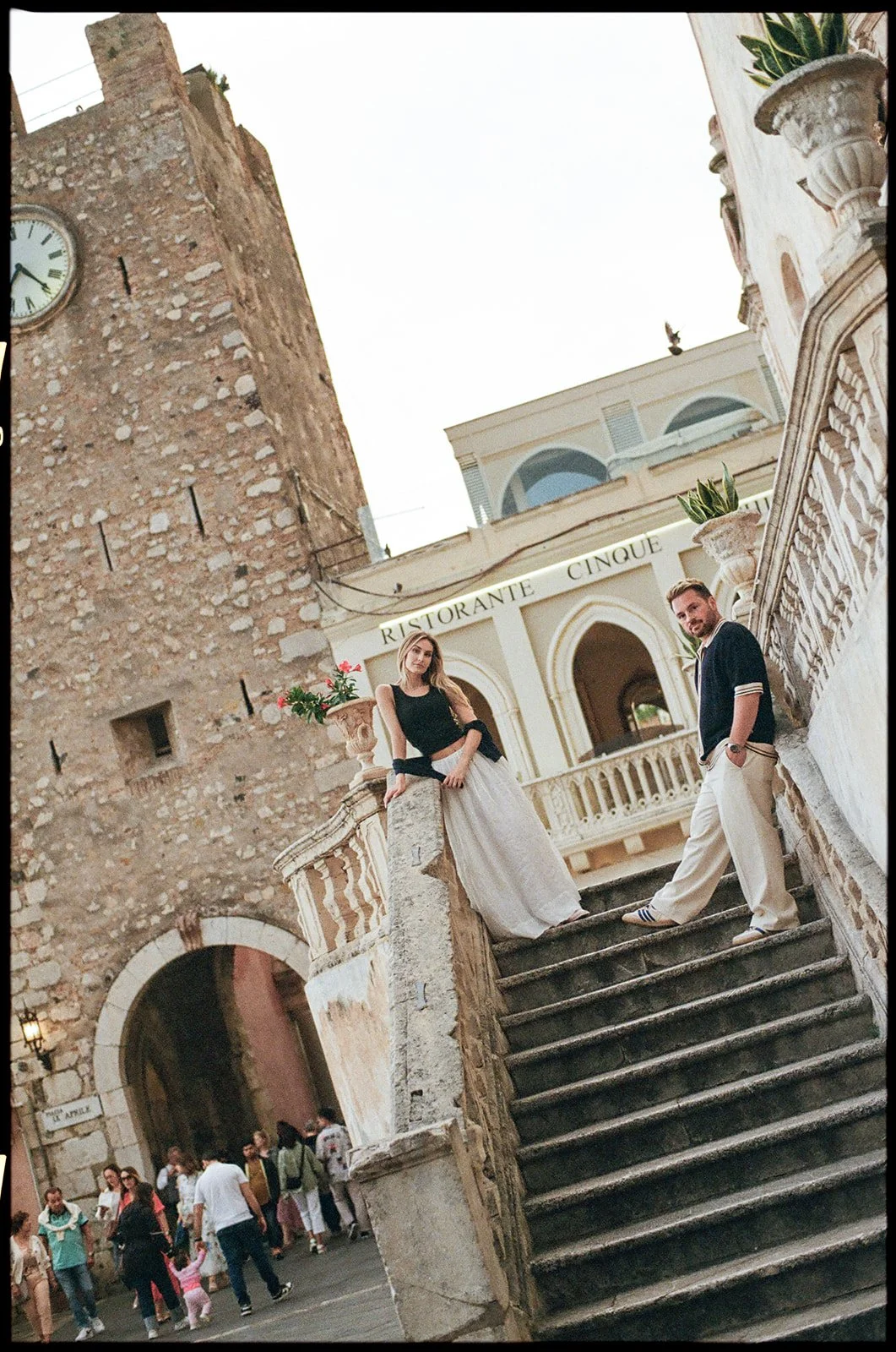 A man and woman stand on stone stairs outside a historic building with a clock tower and an archway, one with their side to the camera, the woman leaning on a stone pillar and the man with hands in pockets, in a lively outdoor setting.