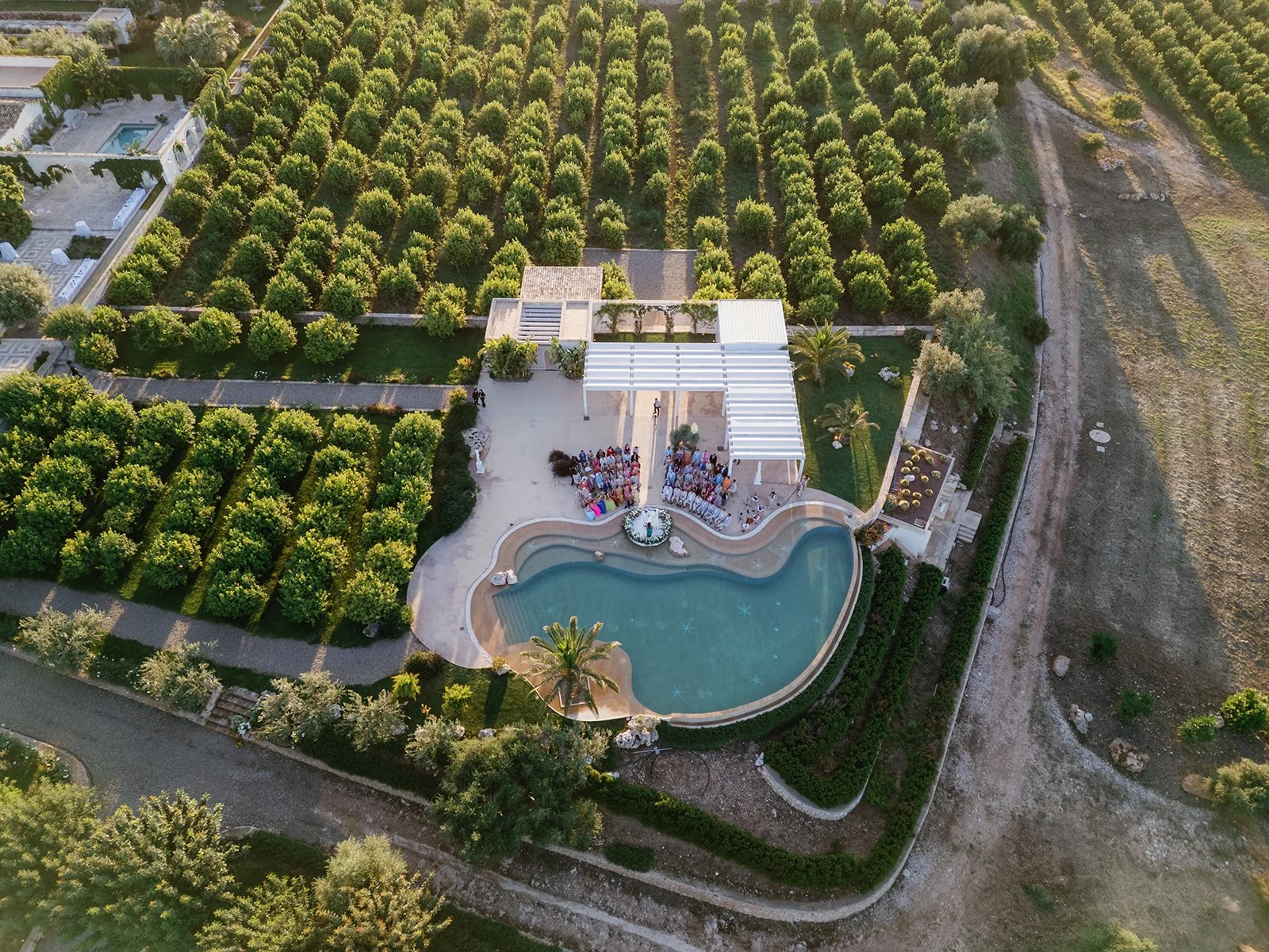 An aerial view of a large outdoor space surrounded by trees and greenery, featuring a swimming pool with a curved shape and patio area with a gathering of people, possibly for an event, with a white pergola and several palm trees nearby.