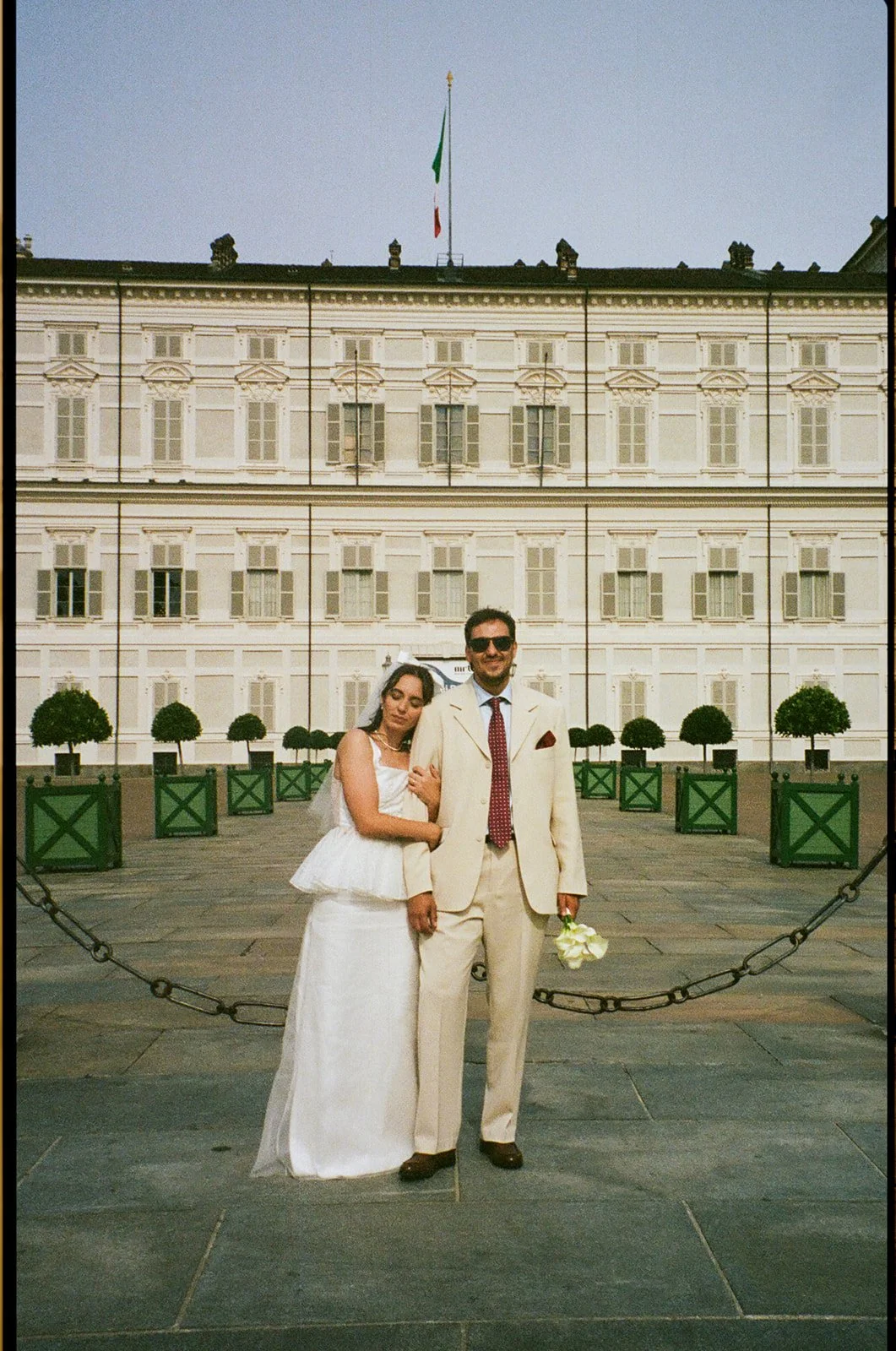 A couple dressed in wedding attire standing in front of a historic building with Italian flag, small trees, and chain barricades.