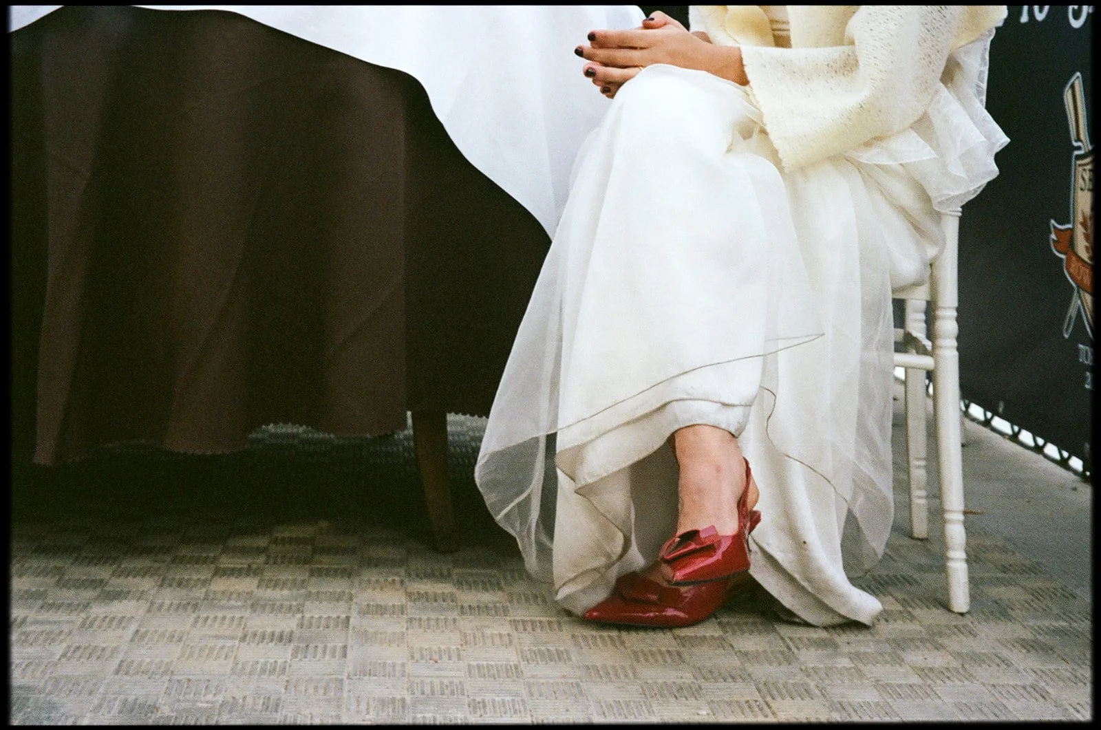 A person sitting on a white chair, wearing a cream-colored sweater, white layered skirt, and red high-heeled shoes, with their hands clasped on their lap, and seated near a black table and a dark crate.
