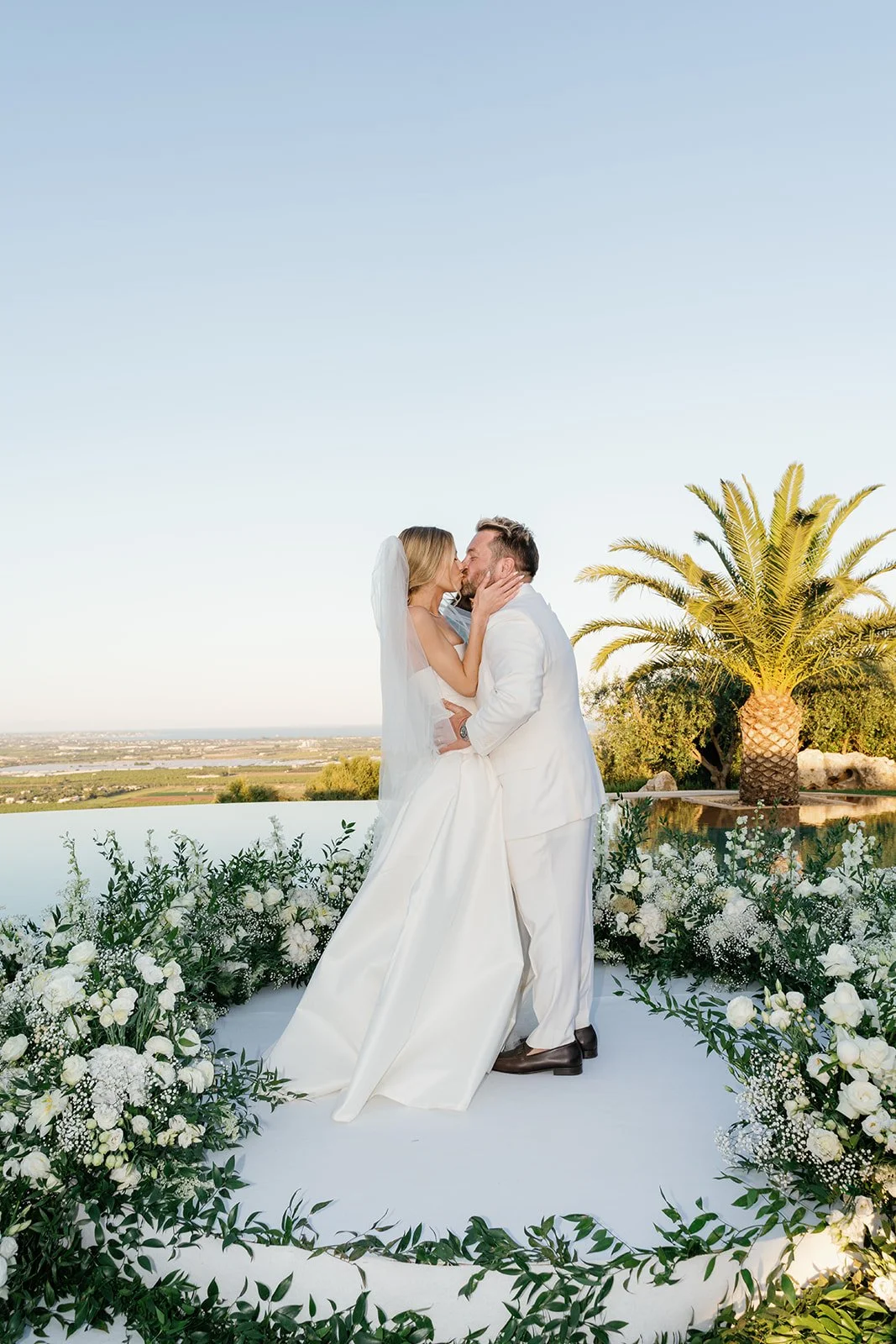 A newlywed couple sharing a kiss outdoors on a wedding day, surrounded by white flowers and greenery, with a view of the landscape and a palm tree in the background.