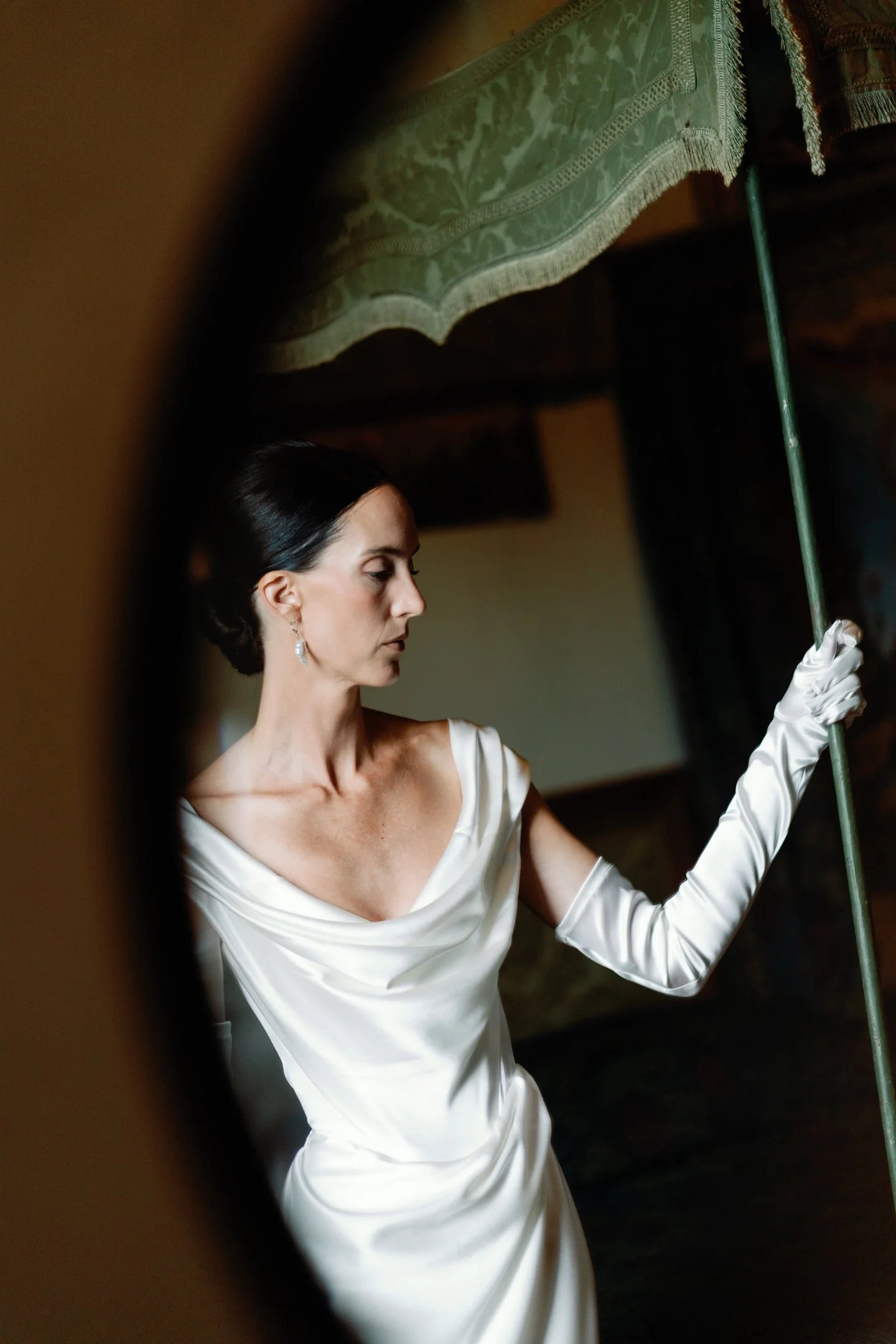 A woman in an elegant white satin dress and long white gloves holding a green umbrella, captured through an oval mirror.