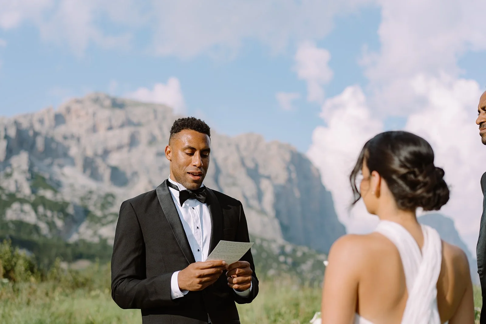 A wedding ceremony outdoors with a man in a tuxedo reading vows, a woman in a white wedding dress, and another man in a suit, set against a mountainous landscape and blue sky with clouds.