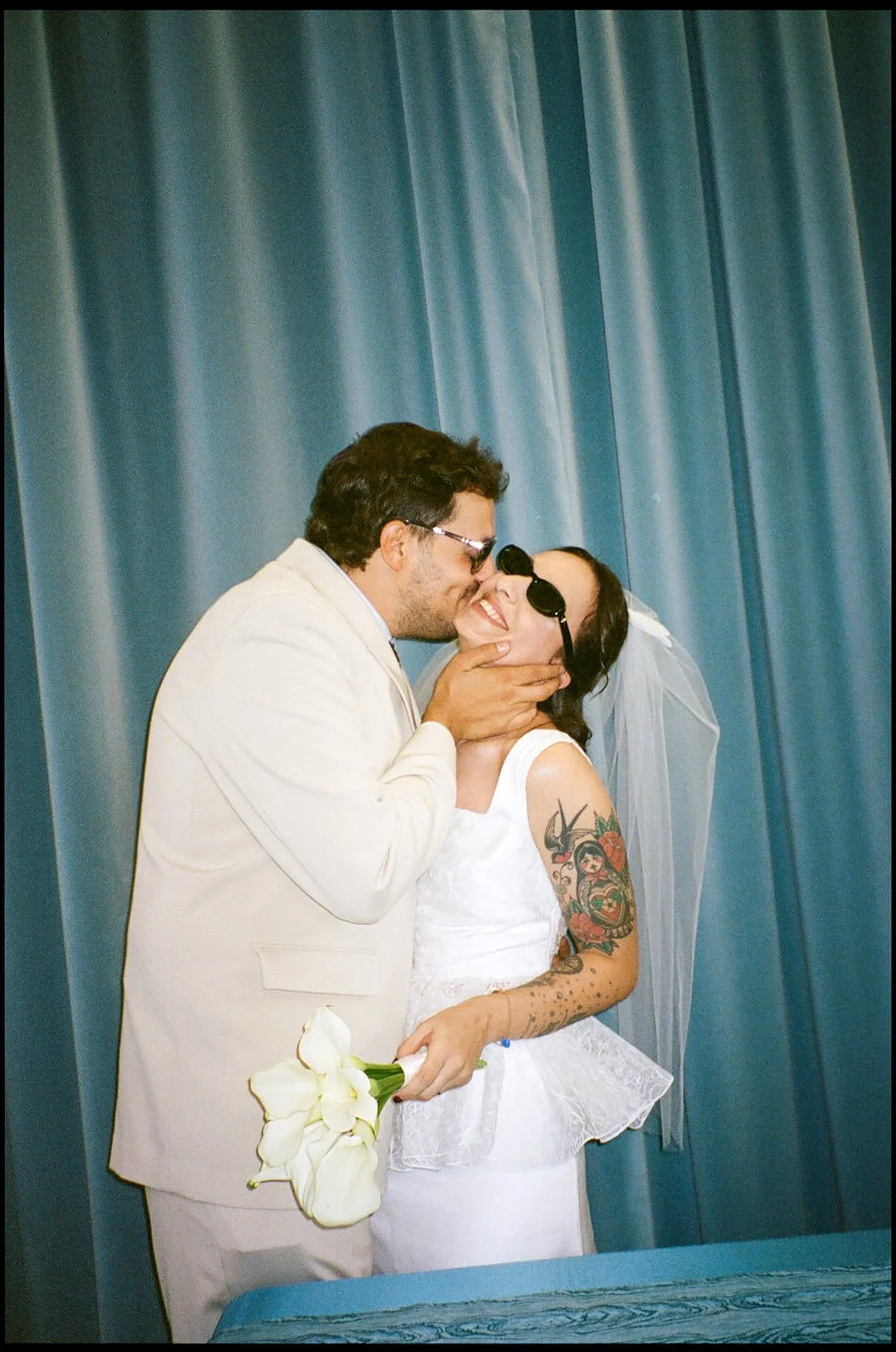 A couple, dressed in wedding attire, sharing an intimate moment with the groom kissing the bride on the lips. The bride wears sunglasses, a white dress, a veil, and holds a bouquet of white calla lilies. The groom wears a light-colored suit. They are standing in front of blue curtains.