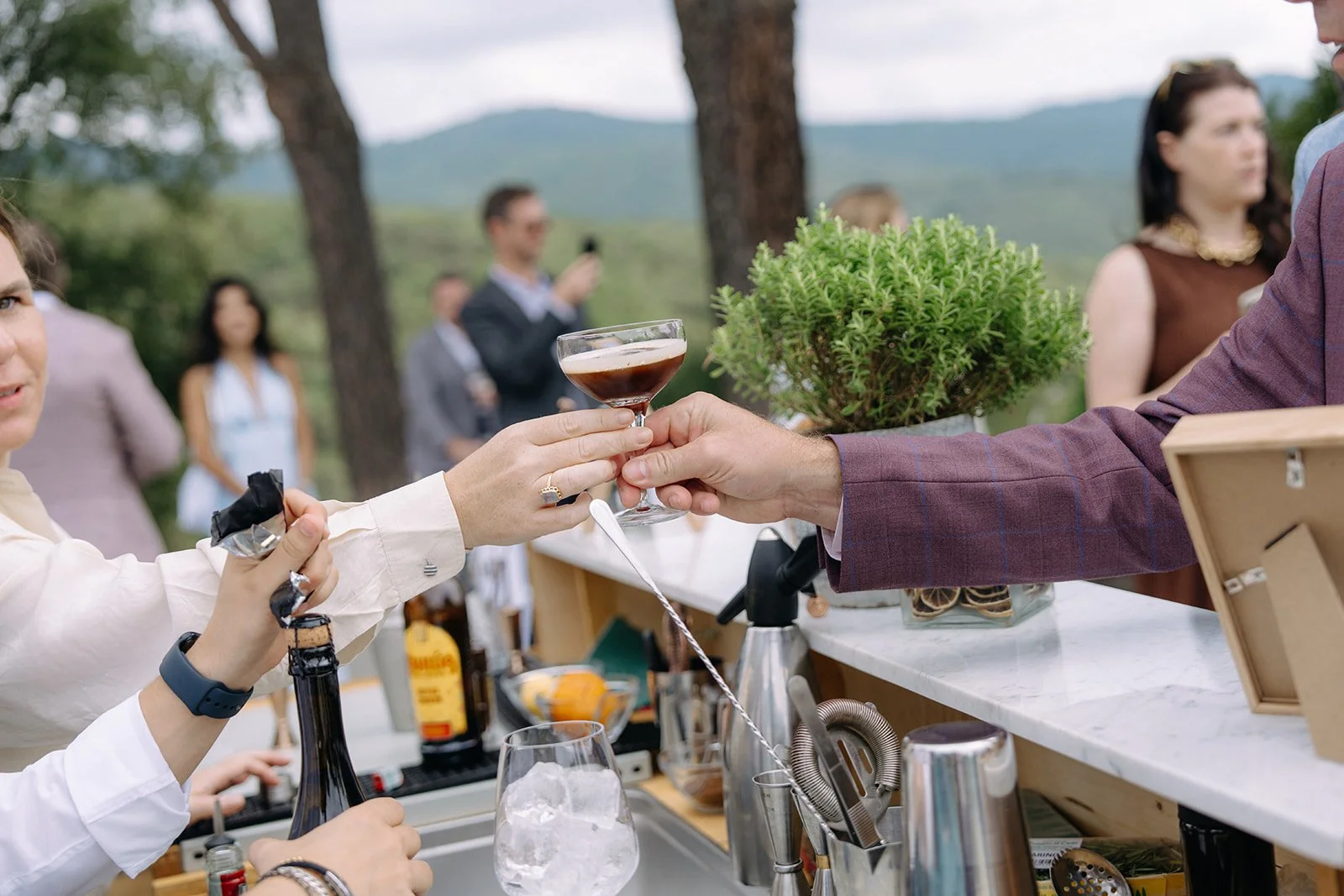 People outdoors at a bar, with a woman in white receiving a cocktail from a man in a burgundy jacket, while other guests mingle in the background.