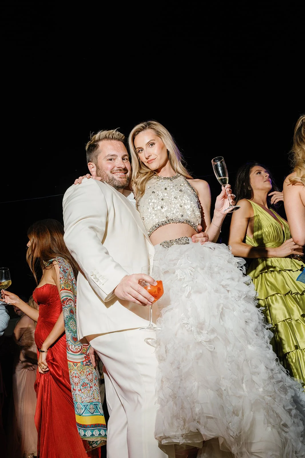Couple at a social event, the man in a white suit holding a cocktail, the woman in a white gown with sequin embellishments and a ruffled skirt, holding a glass of champagne, surrounded by other people in colorful attire.