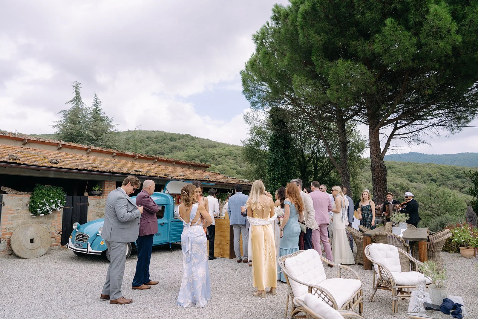 People gathered at an outdoor wedding reception with vintage furniture, food, and drinks, surrounded by greenery and mountains.