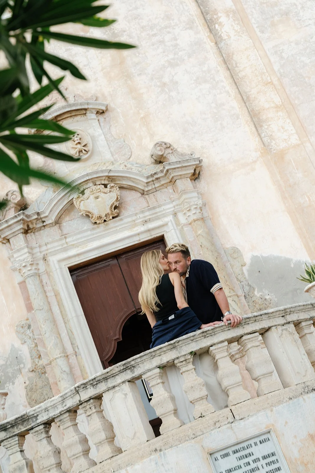 A woman and a man sharing an intimate moment on a stone balcony in front of a historic building with ornate architectural details.