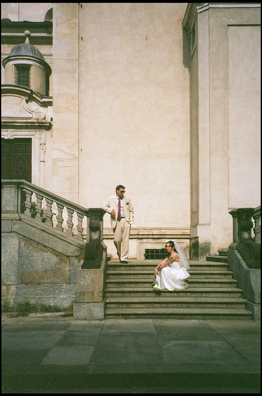 A man and a woman, dressed in formal wedding attire, on the steps outside a historic building with ornate stonework and a small dome. The woman, in a white wedding gown and veil, is sitting on the steps holding flowers, while the man, in a beige suit