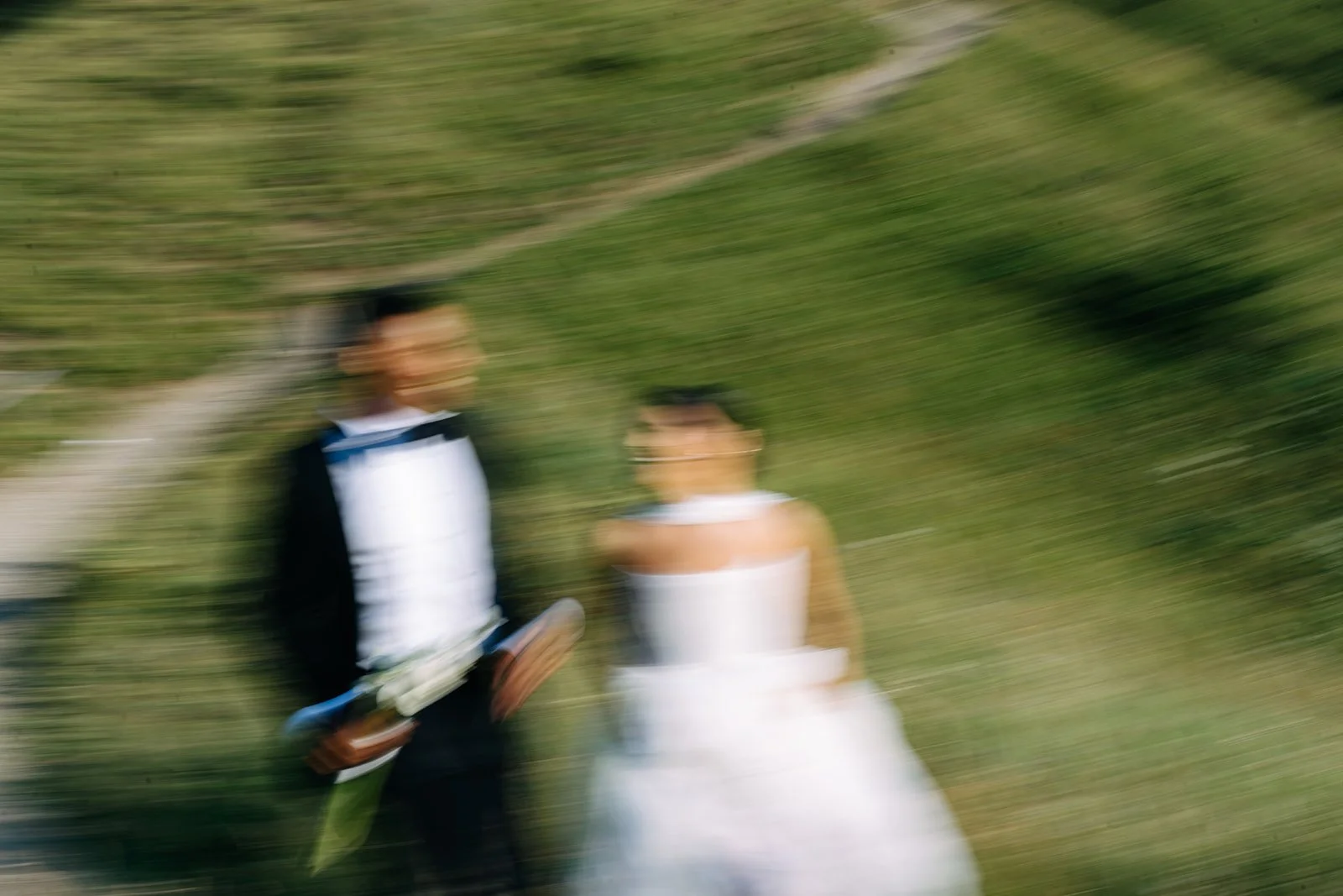 Blurred image of a man in a suit holding a document and a woman in a white dress, standing outdoors on a grassy hill.