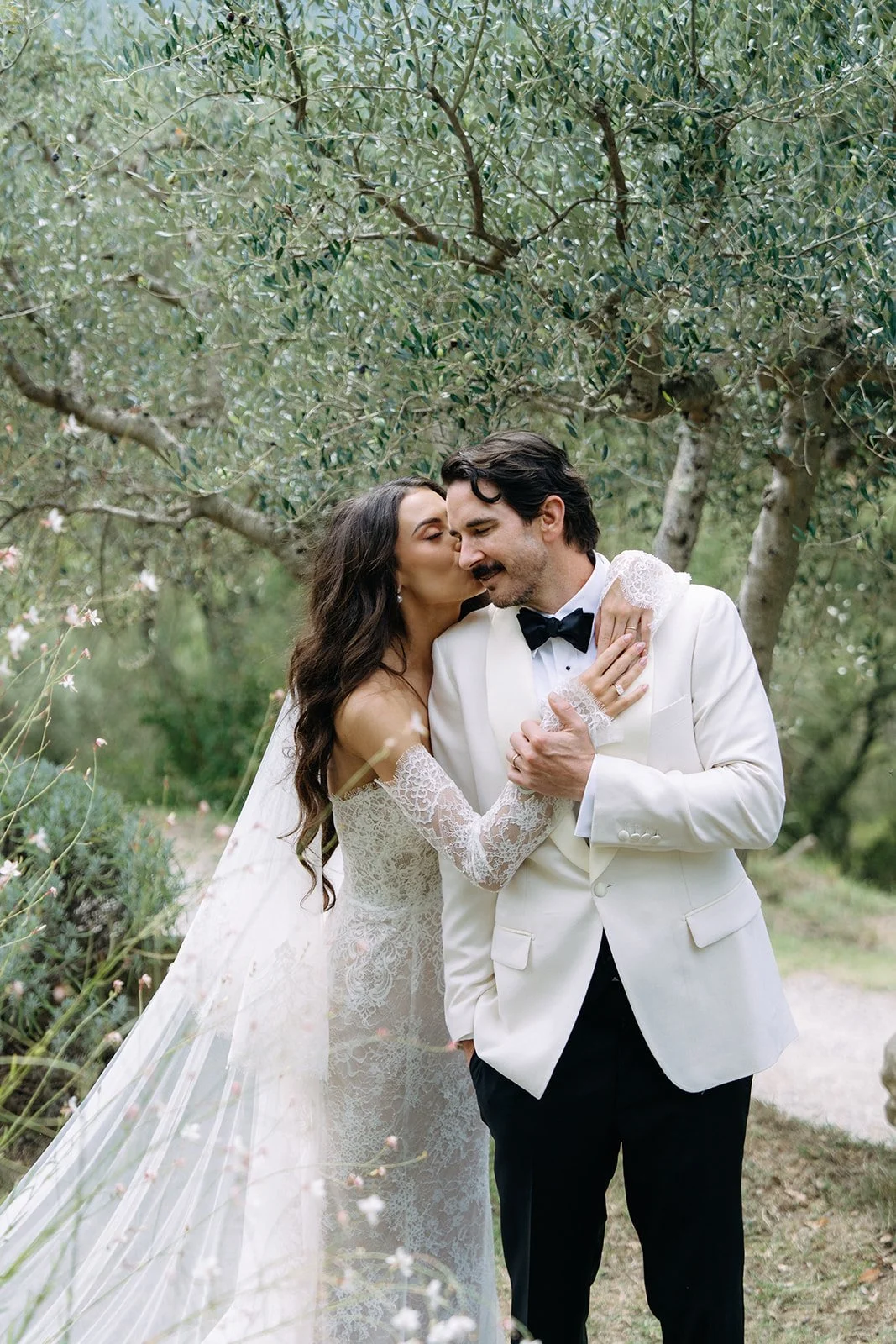 A newlywed couple sharing an intimate moment outdoors, with the bride planting a kiss on the groom's cheek under a tree, both dressed elegantly.