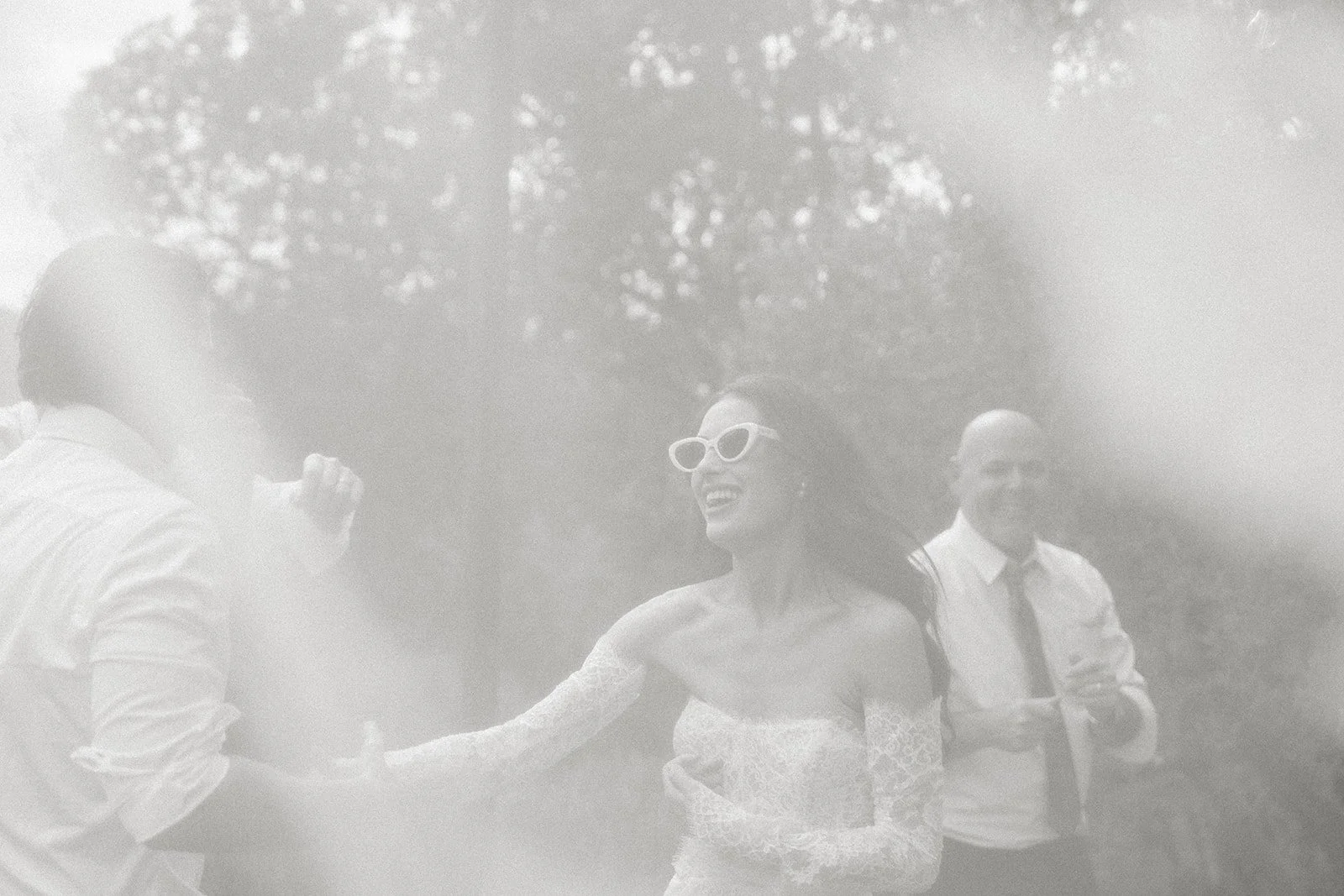 Black and white photo of a woman wearing sunglasses and a lace off-shoulder dress, smiling, as she interacts with a man in a white shirt, outdoors with trees in the background. Another man in a white shirt and tie stands in the background.