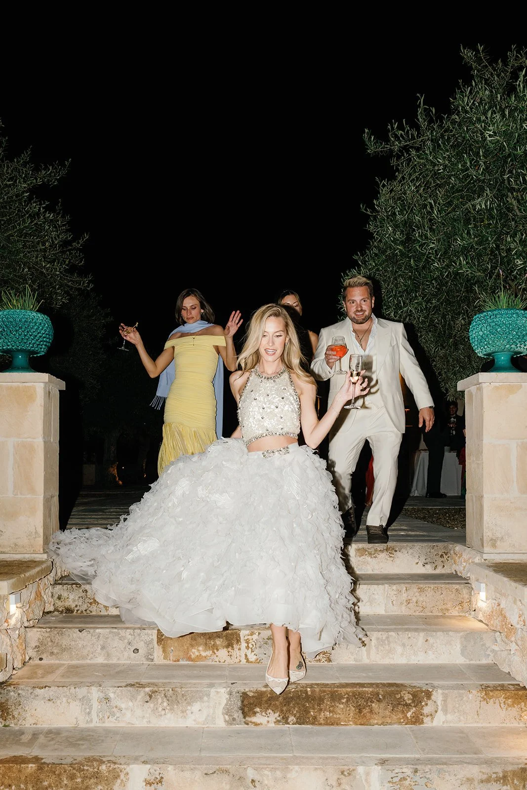 A woman in a white gown and others in formal attire celebrating at night outside, with trees and potted plants in the background.