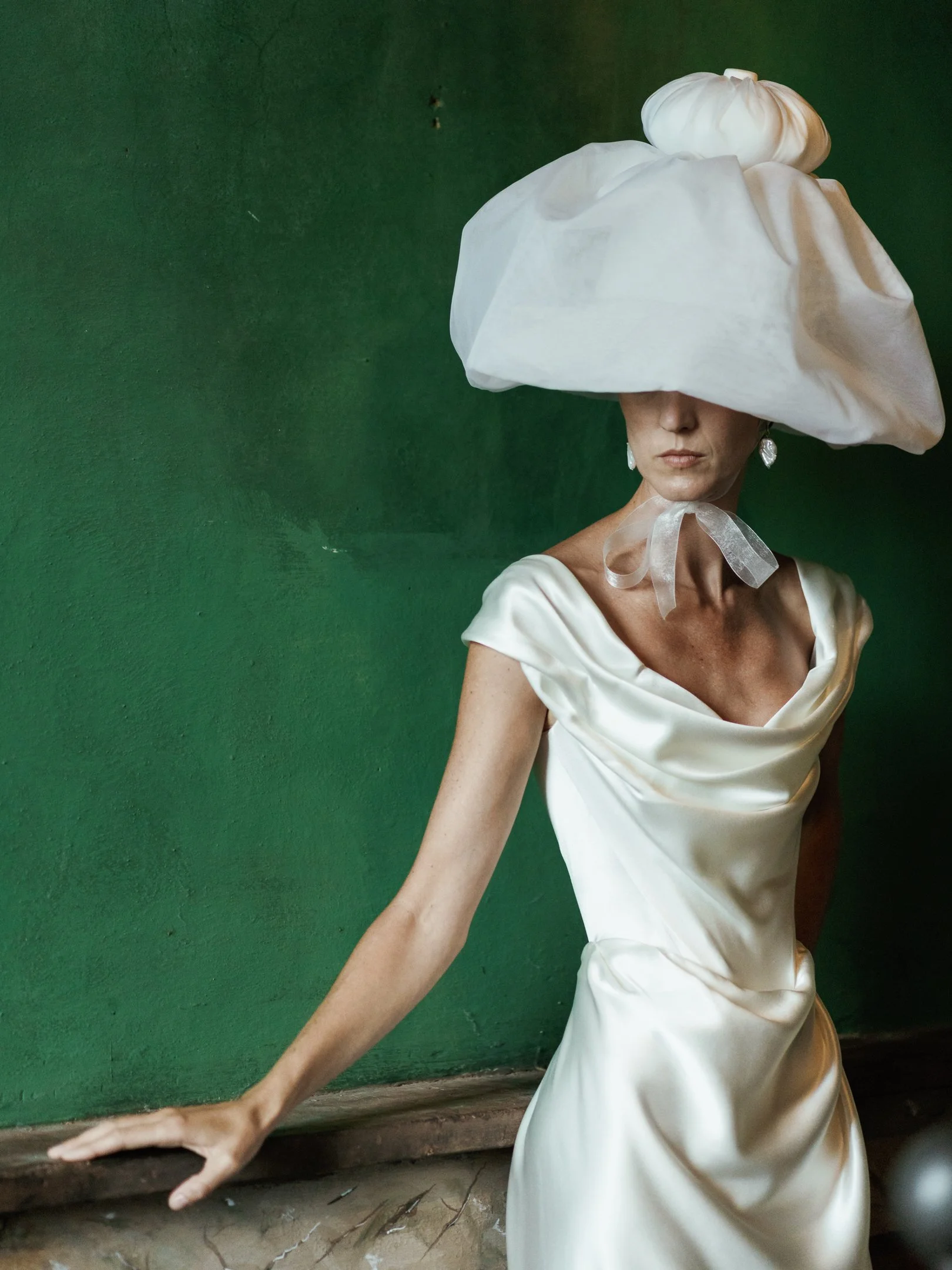 A woman wearing a satin white gown and large white hat with a pumpkin decoration on top, standing against a green wall.
