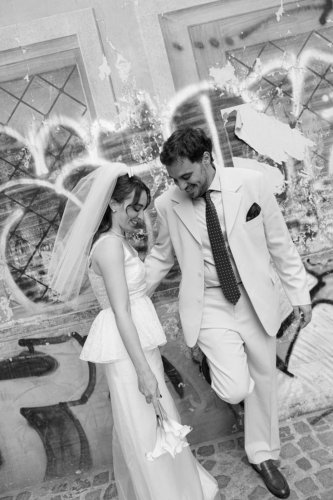 A bride and groom smiling and looking at each other, with the bride holding a bouquet of flowers, standing in front of a graffiti-covered wall.