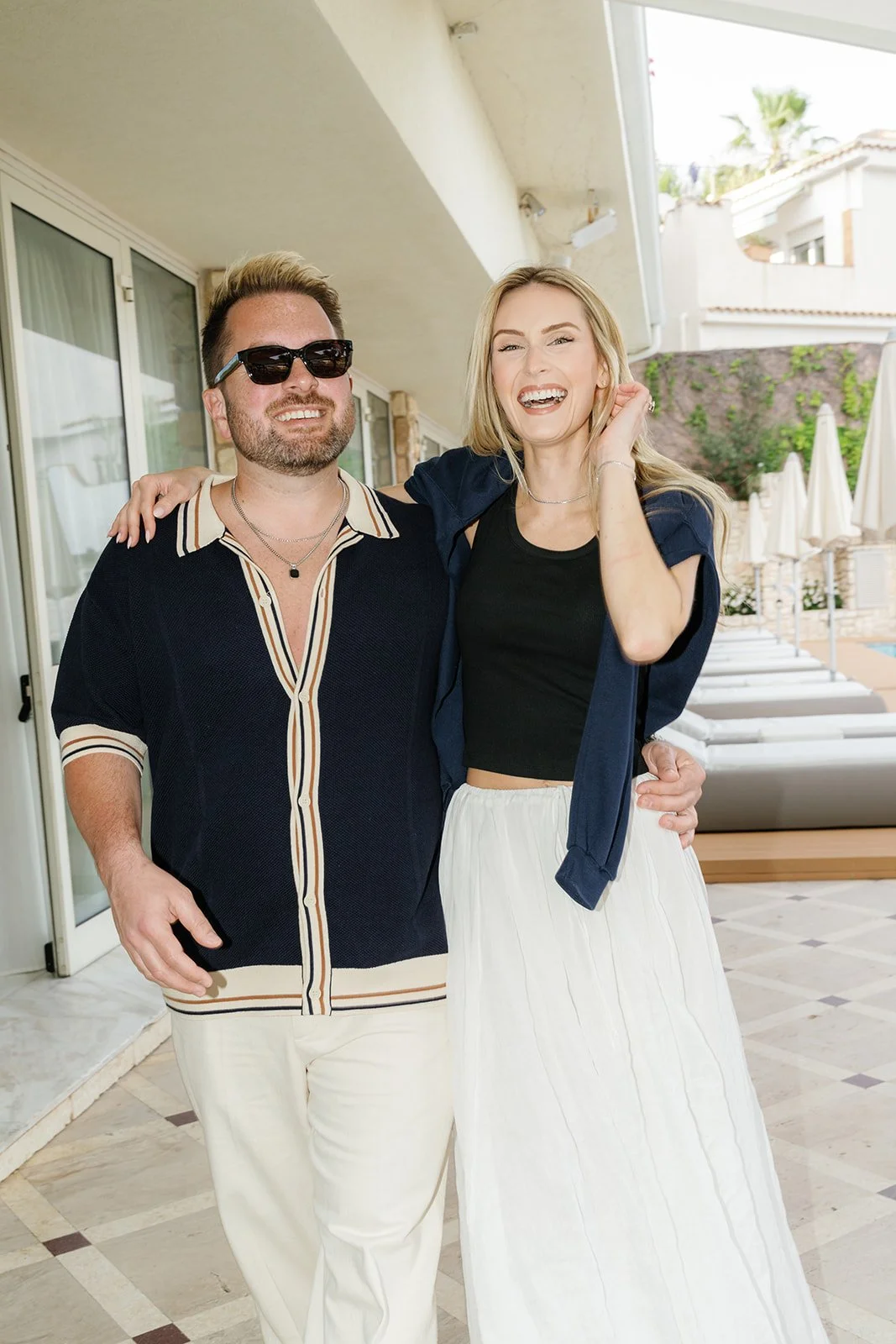 A man and woman smiling and hugging outdoors, with pool chairs and umbrellas in the background.
