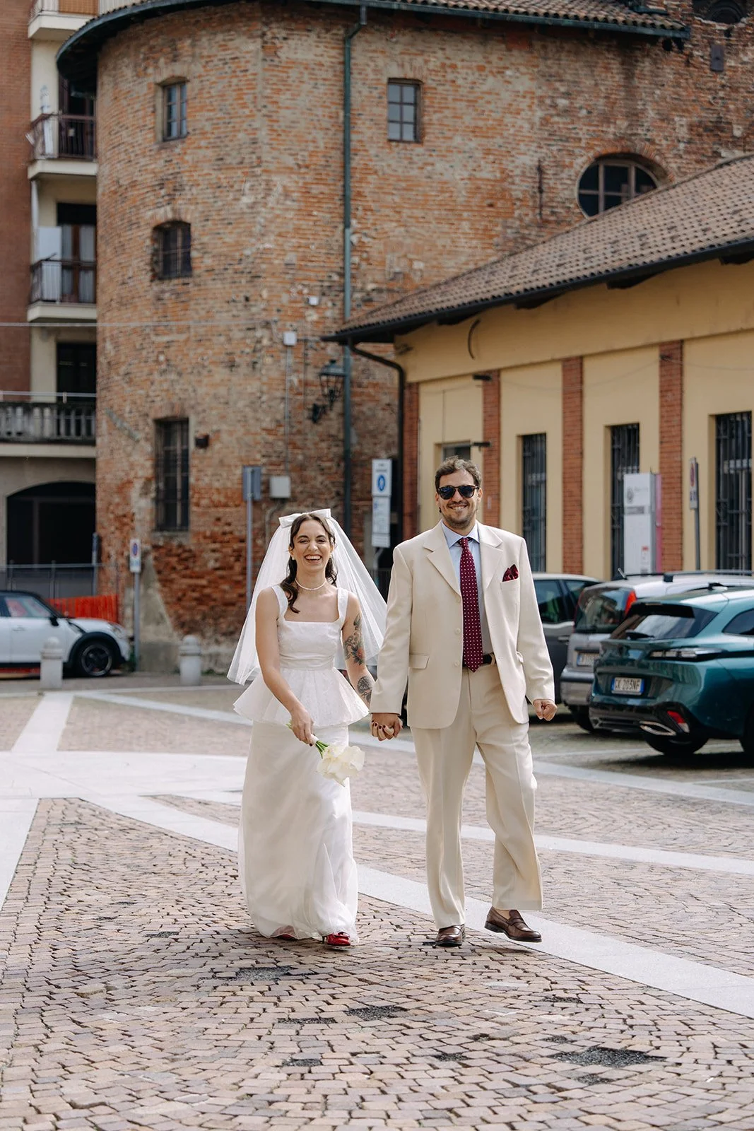 A joyful newlywed couple walking hand in hand on a cobblestone street, with the bride holding a small bouquet of white flowers and wearing a white dress with a veil, while the groom is dressed in a cream suit with a red pocket square and sunglasses, 