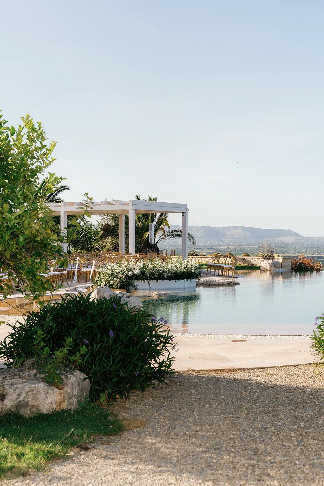 An outdoor event setup near a pool with trees, plants, and mountains in the background.