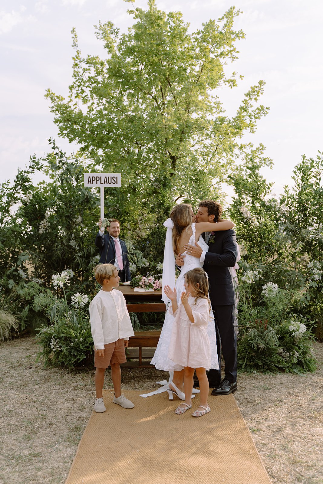A wedding scene outdoors with a bride and groom kissing, surrounded by children and greenery. An officiant in the background holds a sign that says 'APPLAUSI'.