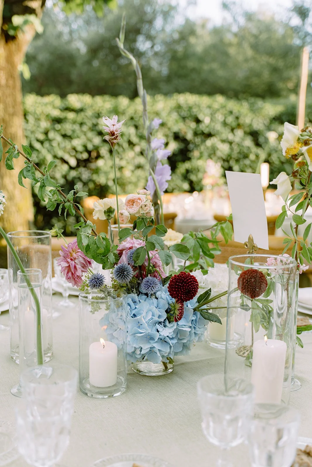 Floral centerpiece with pink, purple, red, and blue flowers on a table with white tablecloth, surrounded by glass candle holders and glasses, outdoors with green trees in the background.