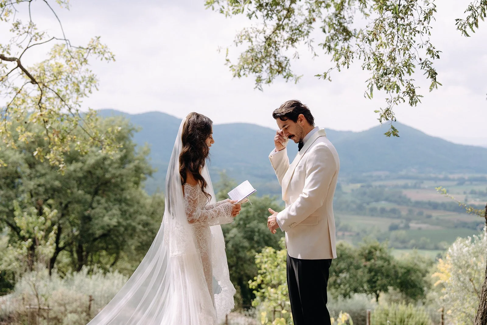 A bride and groom standing outdoors during a wedding ceremony, with mountains and trees in the background. The bride is holding a small book or card and the groom is touching his face, appearing emotional.