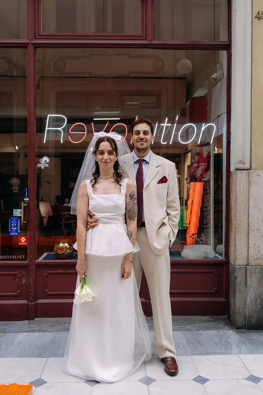 A bride and groom posing in front of a store window; she is holding flowers, he is dressed in a cream suit, standing inside a store with a neon sign that says 'Revolution' in the background.