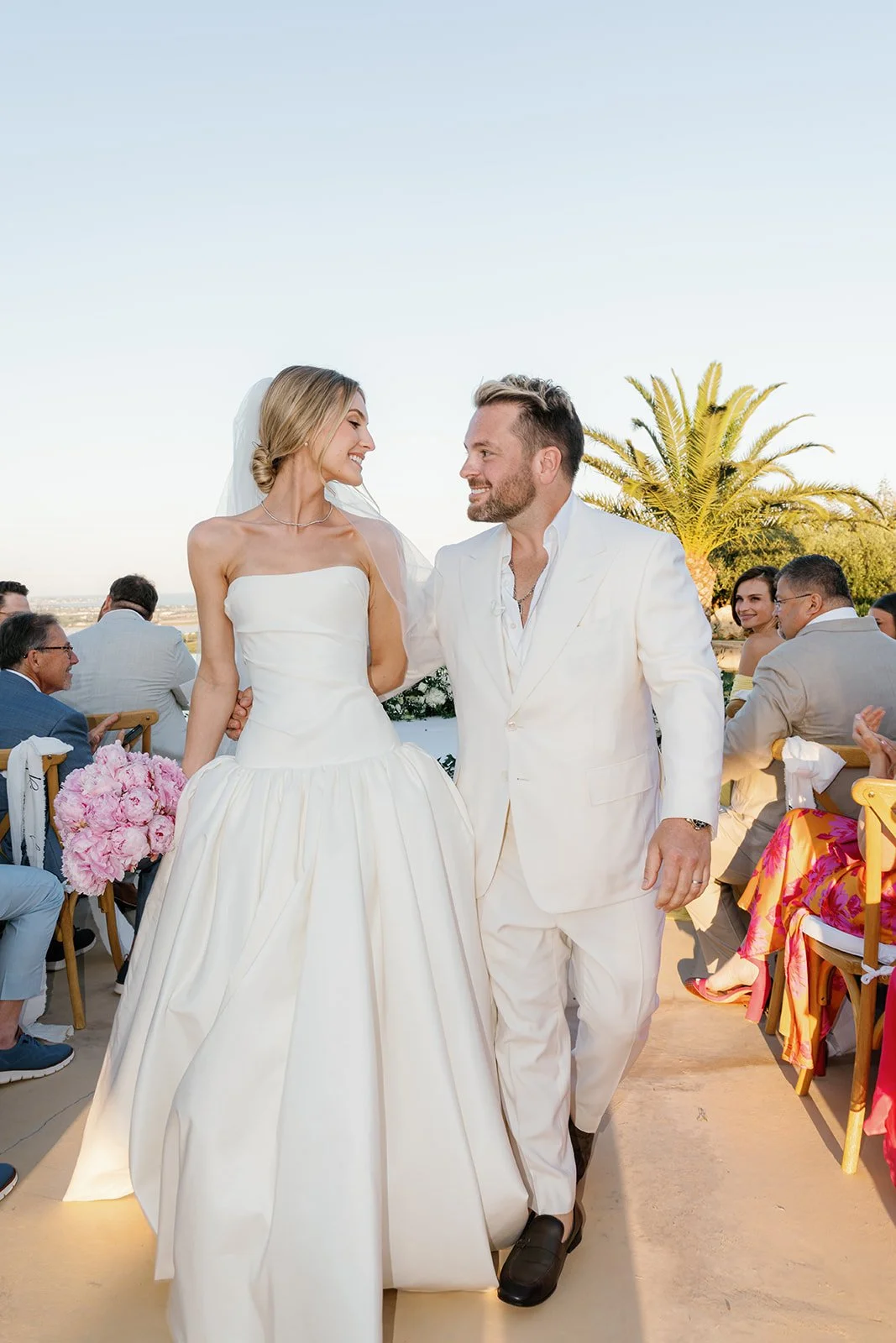 Bride and groom smiling at each other during their outdoor wedding ceremony with guests seated nearby and a palm tree in the background.