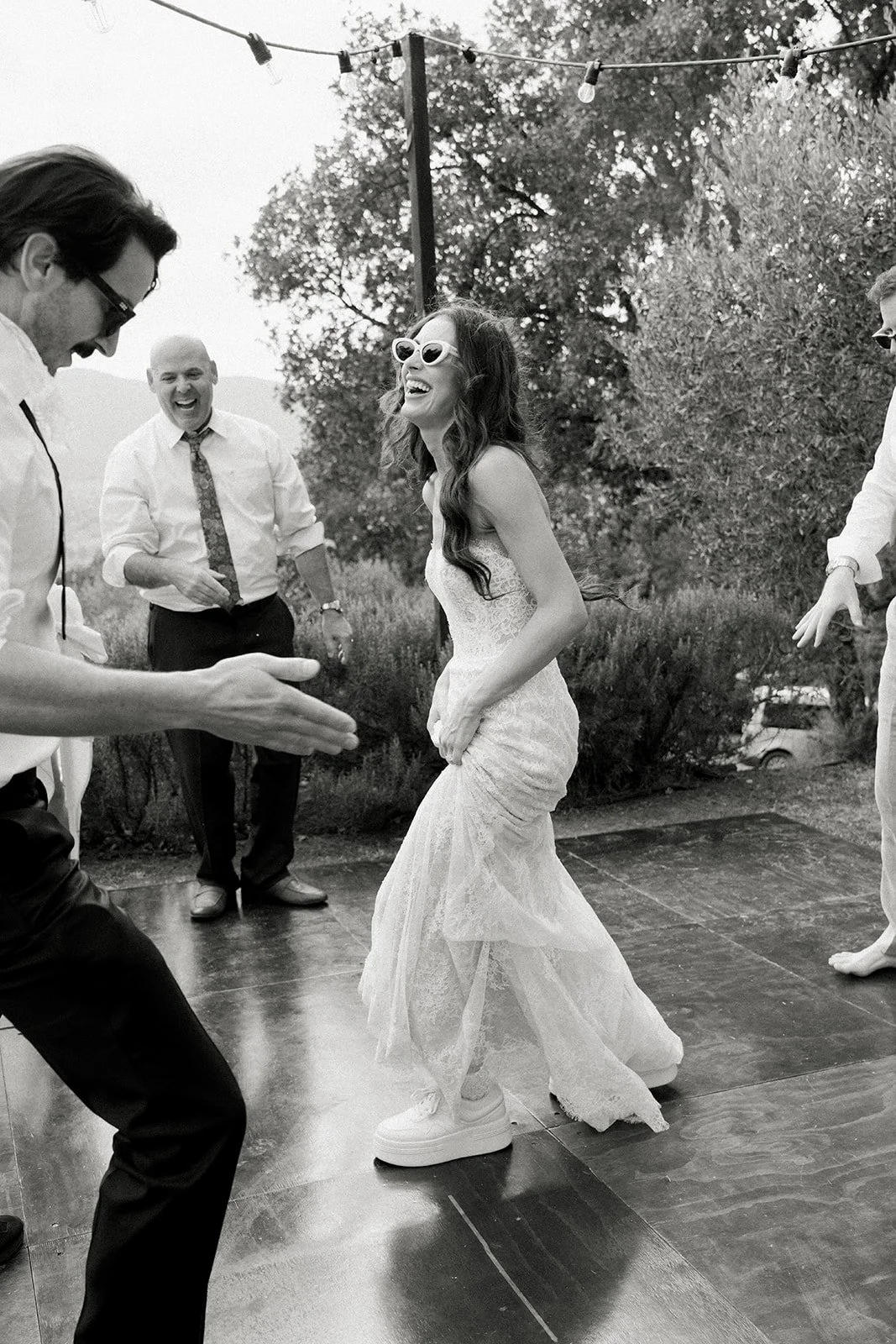A woman dancing and laughing with friends outdoors, wearing sunglasses and a lace dress, with others clapping and smiling nearby.