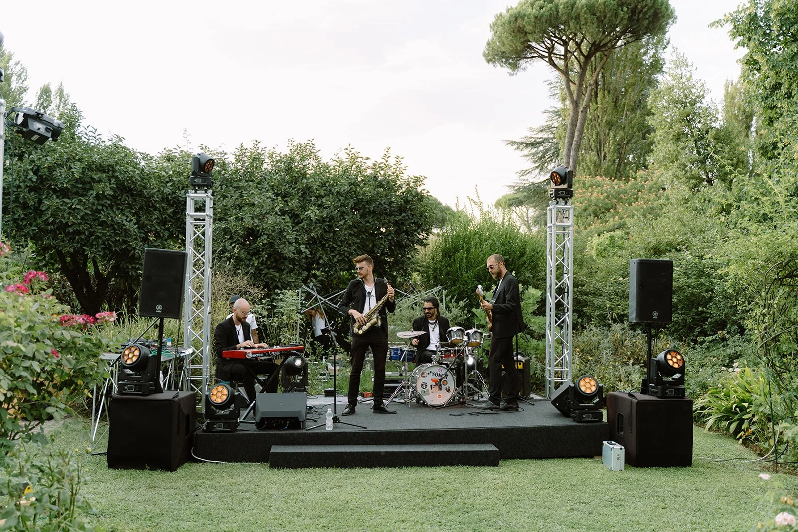A band of four musicians performing outdoors on a small stage surrounded by greenery and trees, with stage lighting and sound equipment.