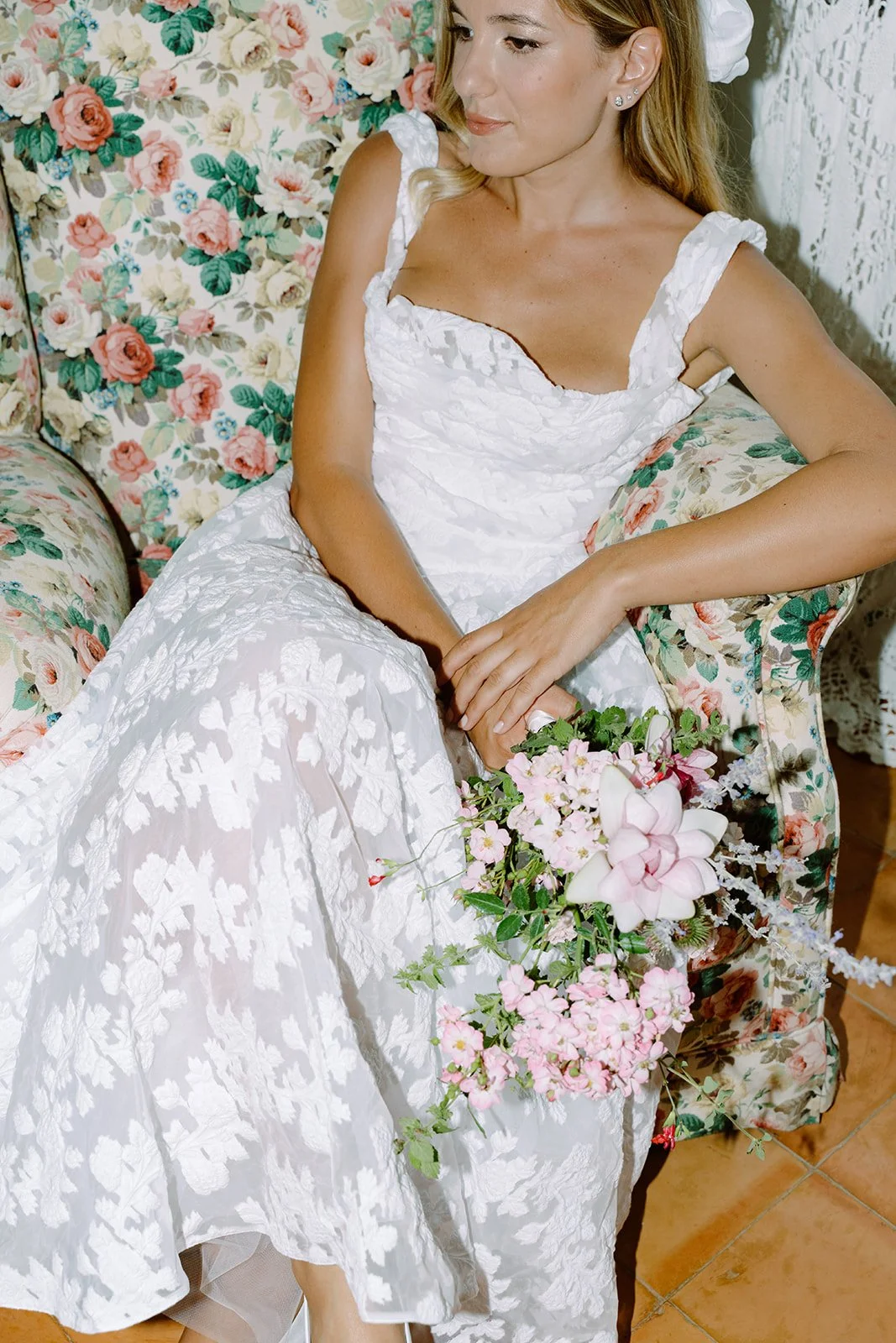 A woman in a white floral dress sitting on a floral-patterned sofa, holding a bouquet of pink and white flowers.