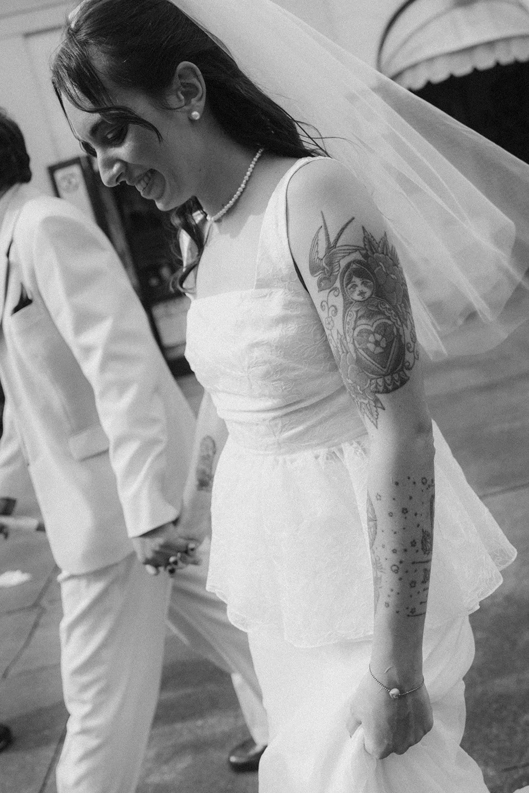 A woman in a white wedding dress with tattoos on her arm, smiling and holding hands with someone in a white suit during a wedding ceremony.