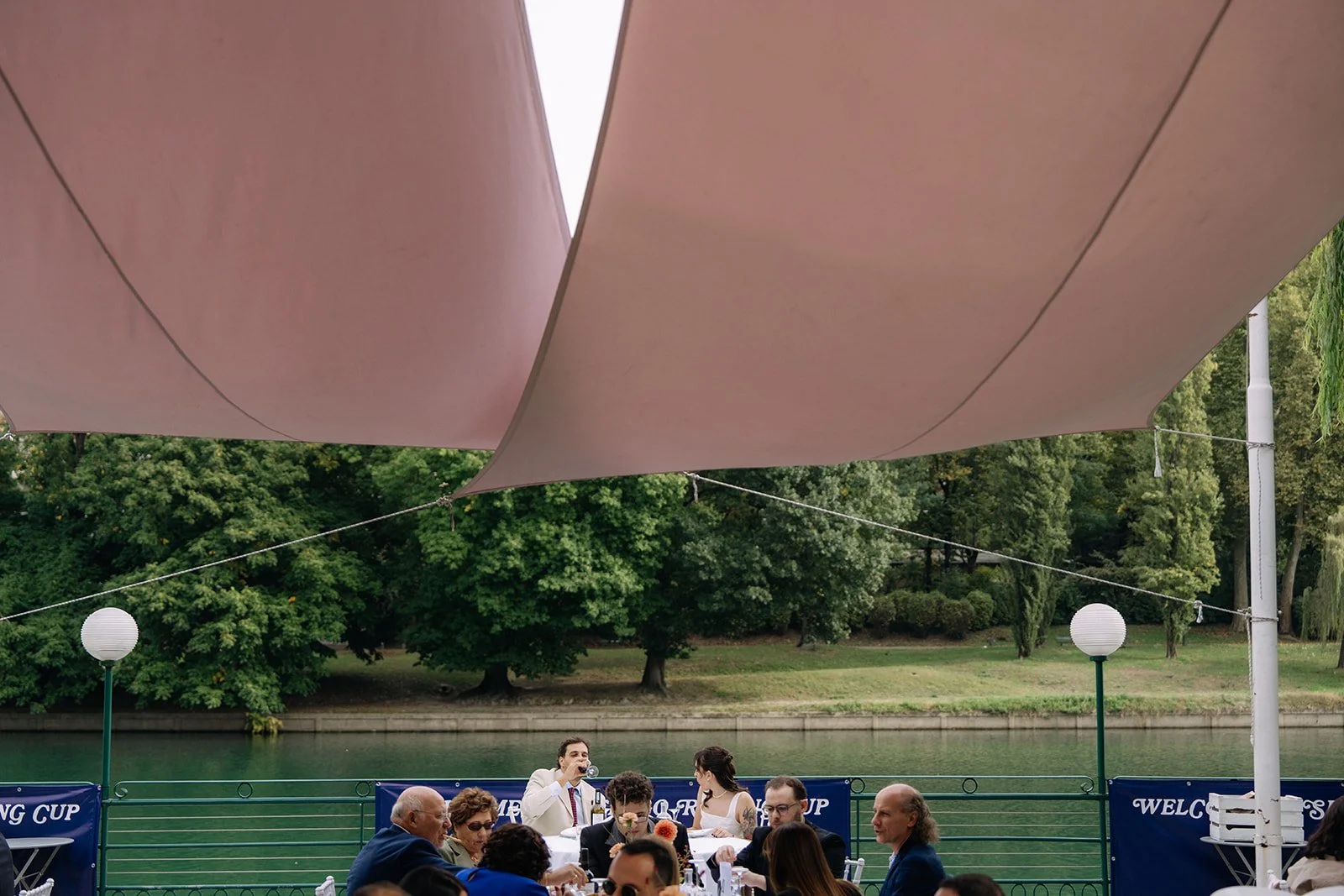 People dining outdoors by a river, shaded by pink fabric overhead, with greenery and trees in the background.