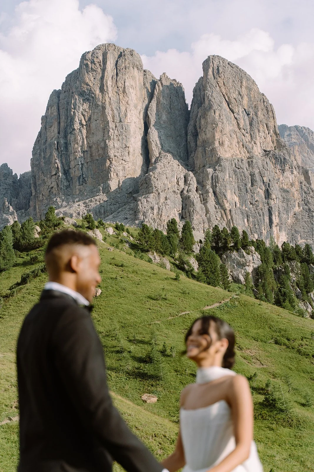 Blurred couple in formal attire holding hands outdoors with tall rocky mountains and green hillside in the background.