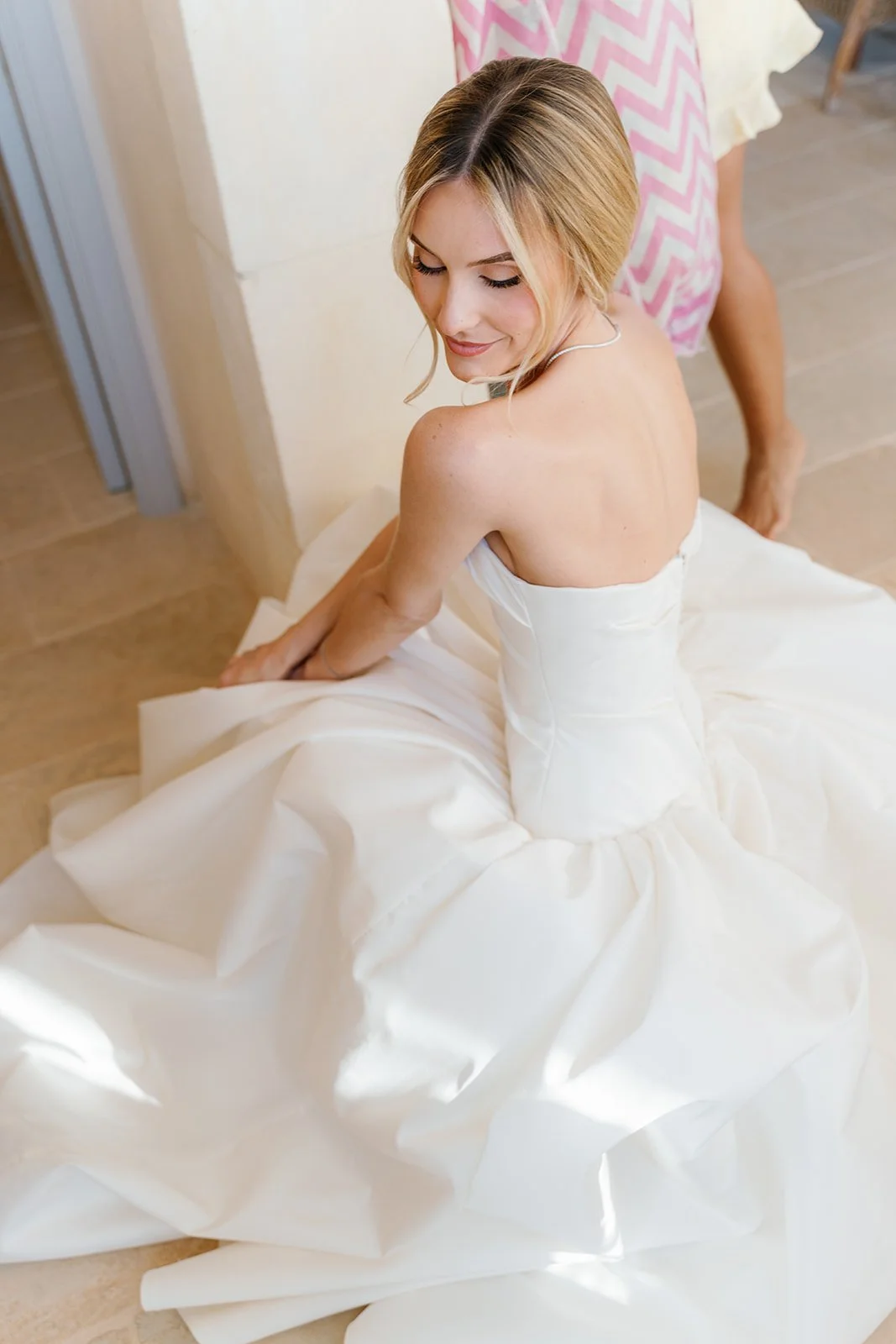 A woman in a white wedding dress sitting on the floor, smiling with her eyes closed, near a beige wall and wooden floor, with another person in a pink and white patterned dress in the background.