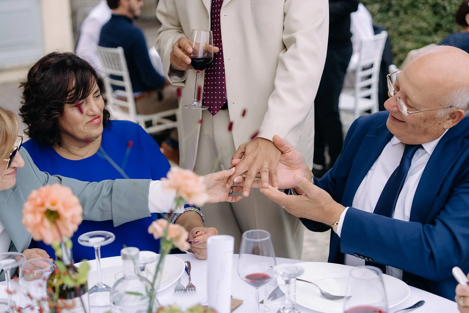 A group of people at a social gathering, with an older man in a blue suit and glasses placing a ring on a woman's finger, others looking on and smiling, with wine glasses and flowers on the table.