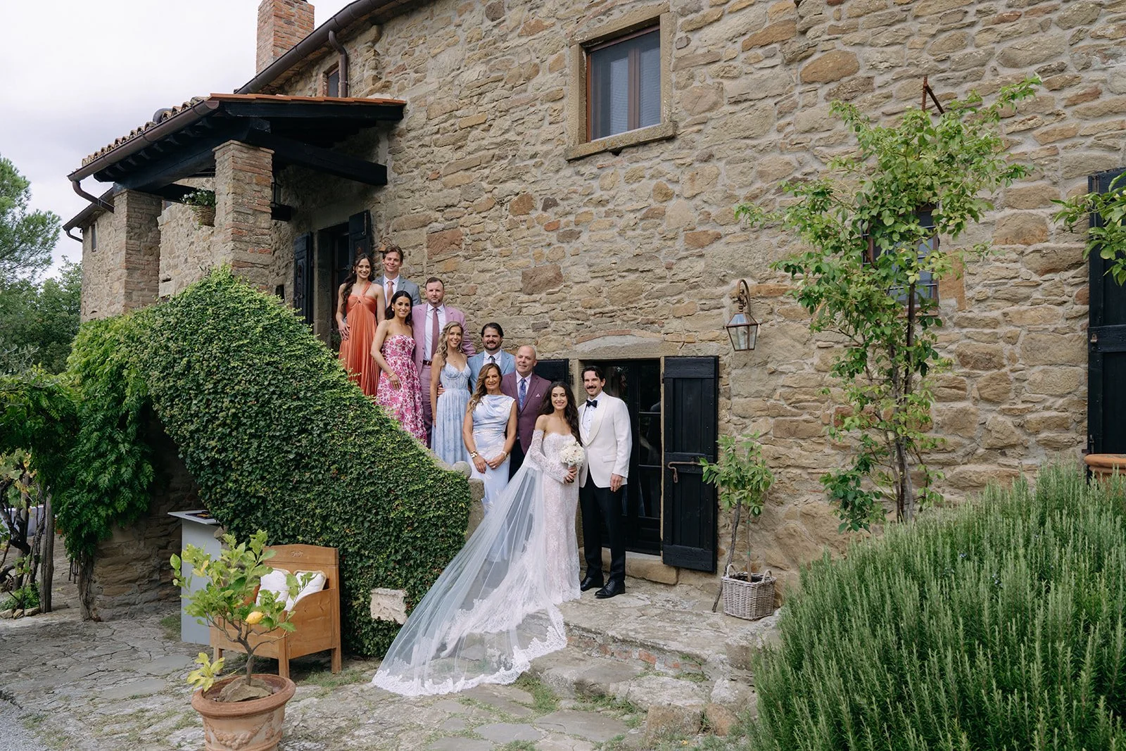 A wedding group photo taken outside a rustic stone building featuring the bride and groom in front, with the bride wearing a white wedding gown and veil, and the groom in a white tuxedo jacket. Behind them are bridesmaids and groomsmen dressed in col