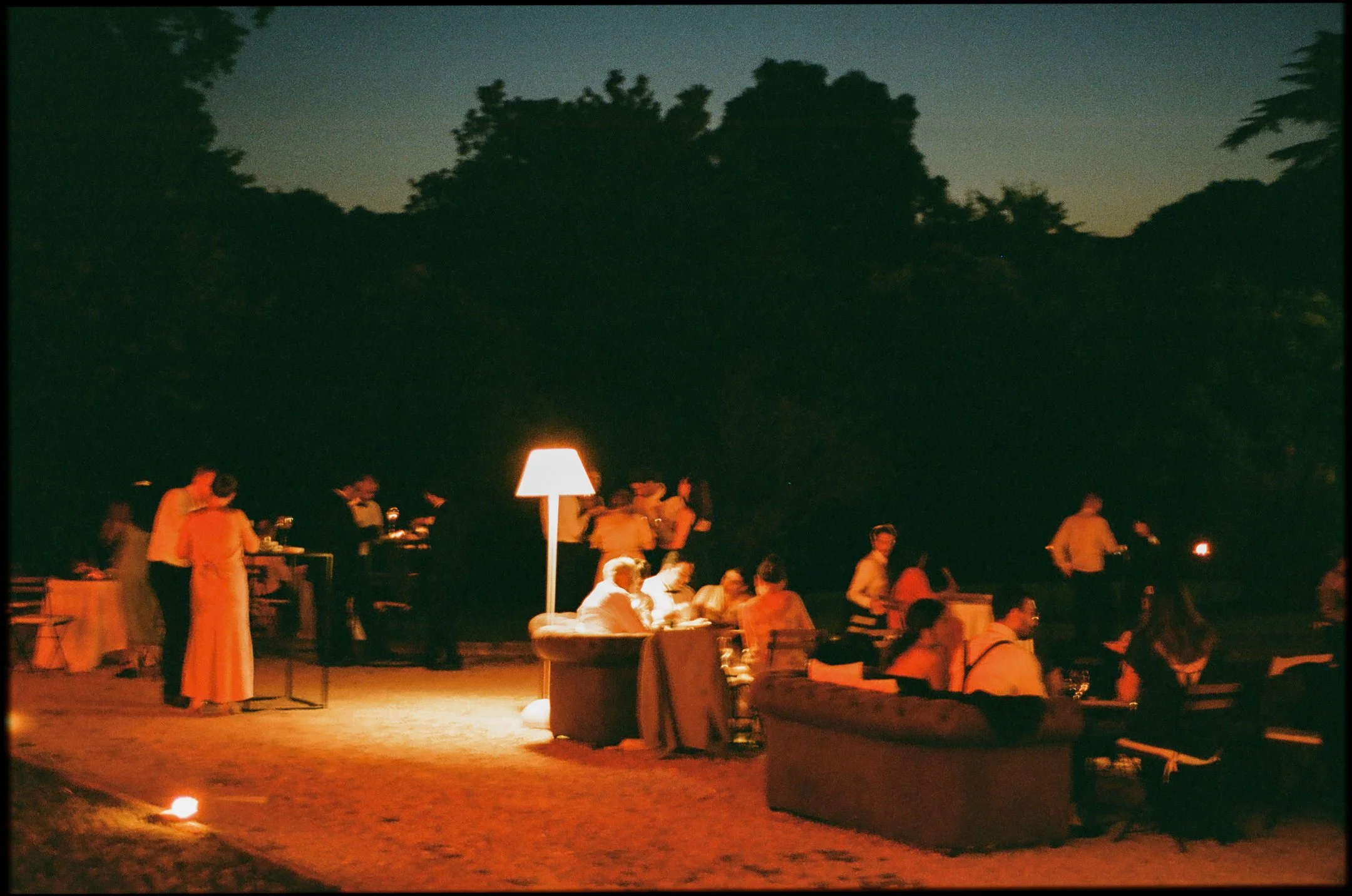 People gathered outdoors at night, sitting on couches and standing, illuminated by a lamp and candles, with trees in the background.