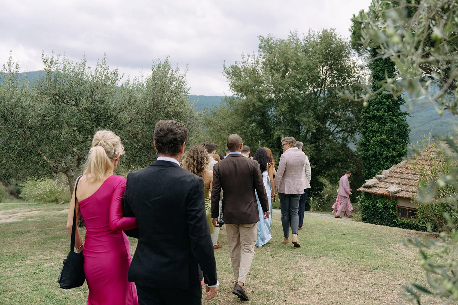 A group of people dressed in formal and semi-formal attire, walking outdoors on a grassy area with trees and mountains in the background, heading towards a small building with a tiled roof.