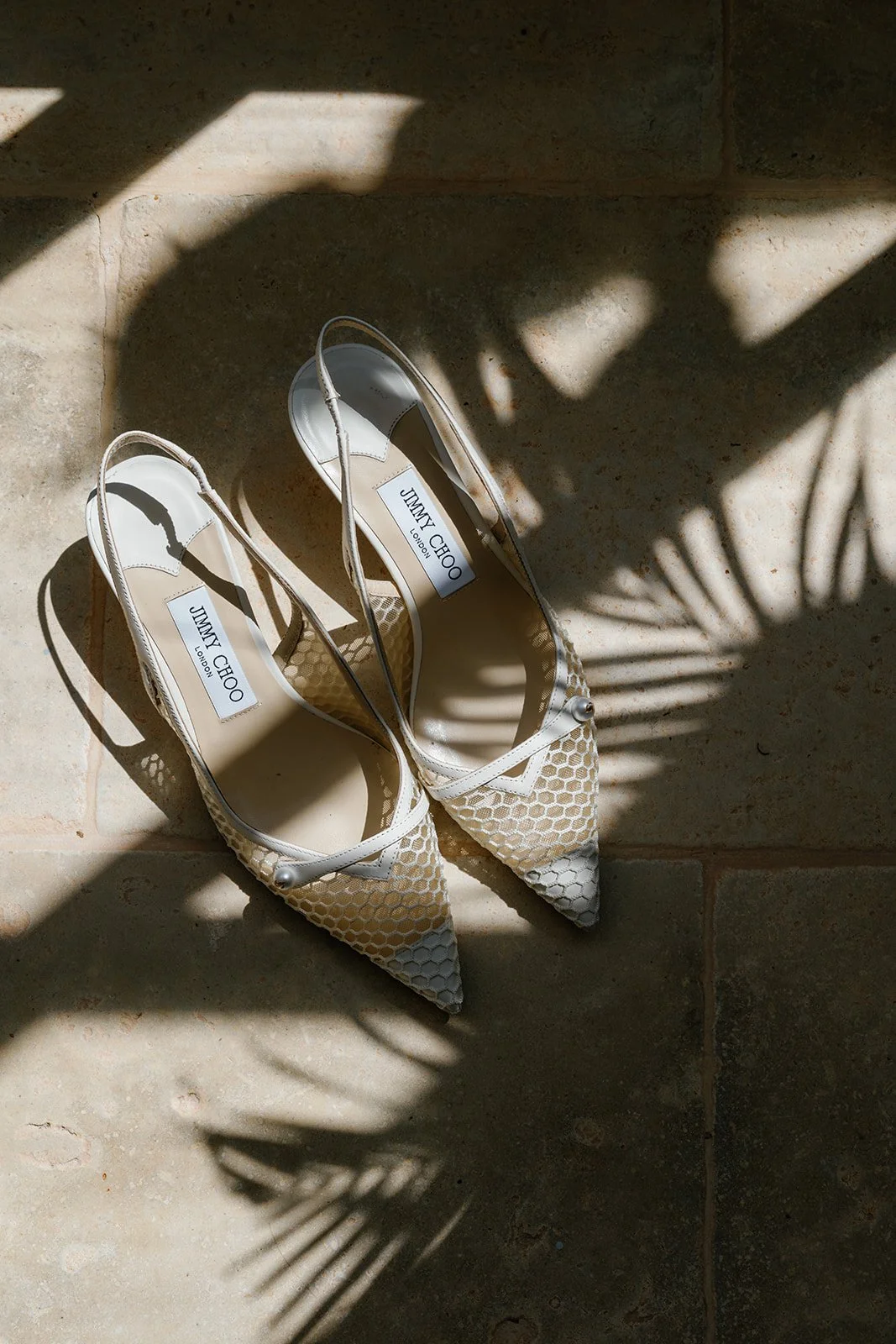 A pair of cream and beige pointed-toe Jimmy Choo high heels with thin ankle straps, placed on a tiled floor. Shadows of a plant are cast across the shoes, creating a pattern on the floor.